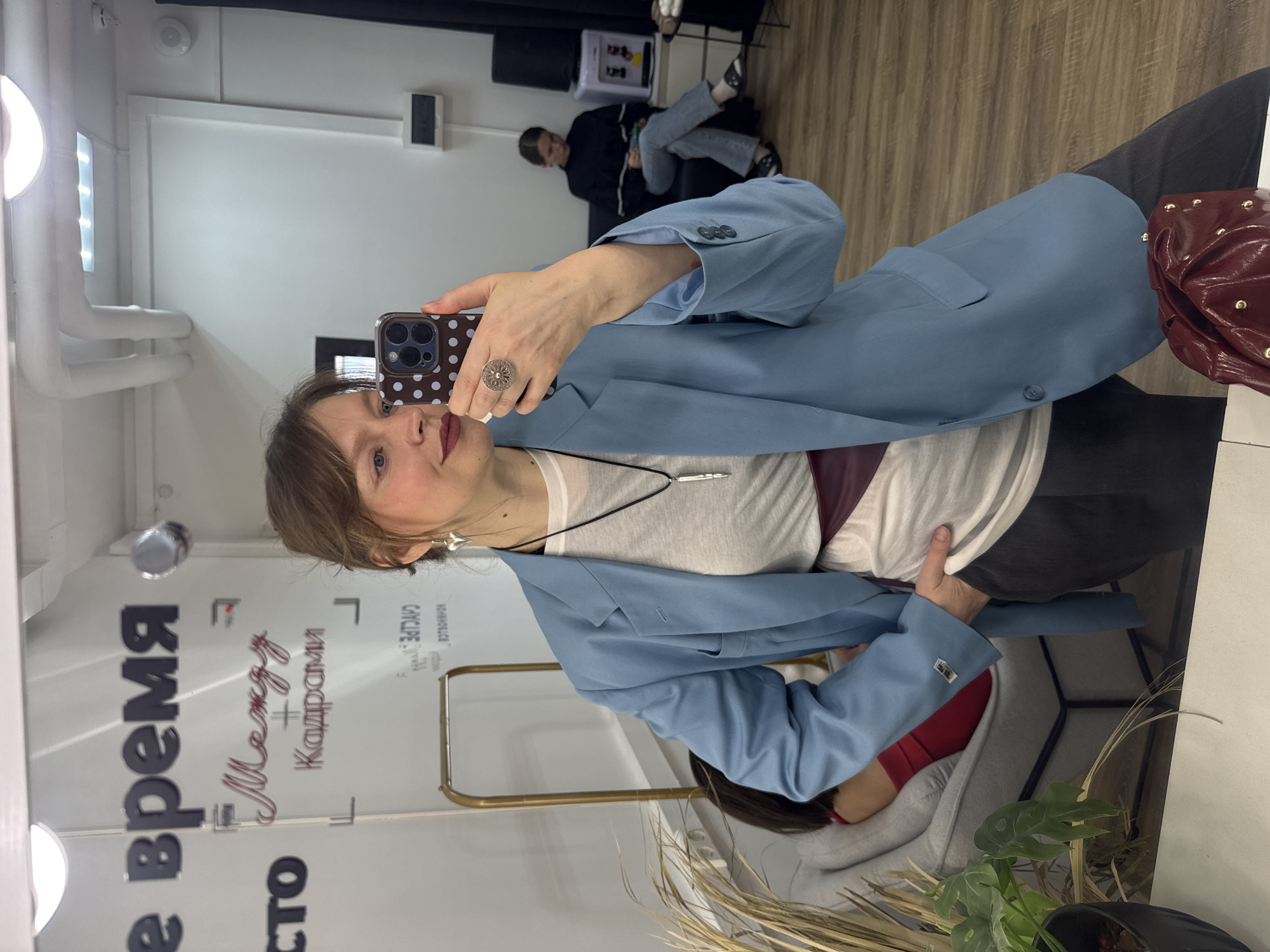 A woman is taking a selfie in a mirror. She is wearing a light blue blazer over a white t-shirt, with a silver pendant necklace. She has a ring on her finger and is holding a smartphone with a polka dot case. Behind her, a person is seated on a chair. The environment appears to be a store, with text in Russian ("время между кадрами") visible on the mirror, possibly indicating "Time Between Frames." The location is Izhevsk, Russia. Other details include clothing racks, a plant, and furniture.