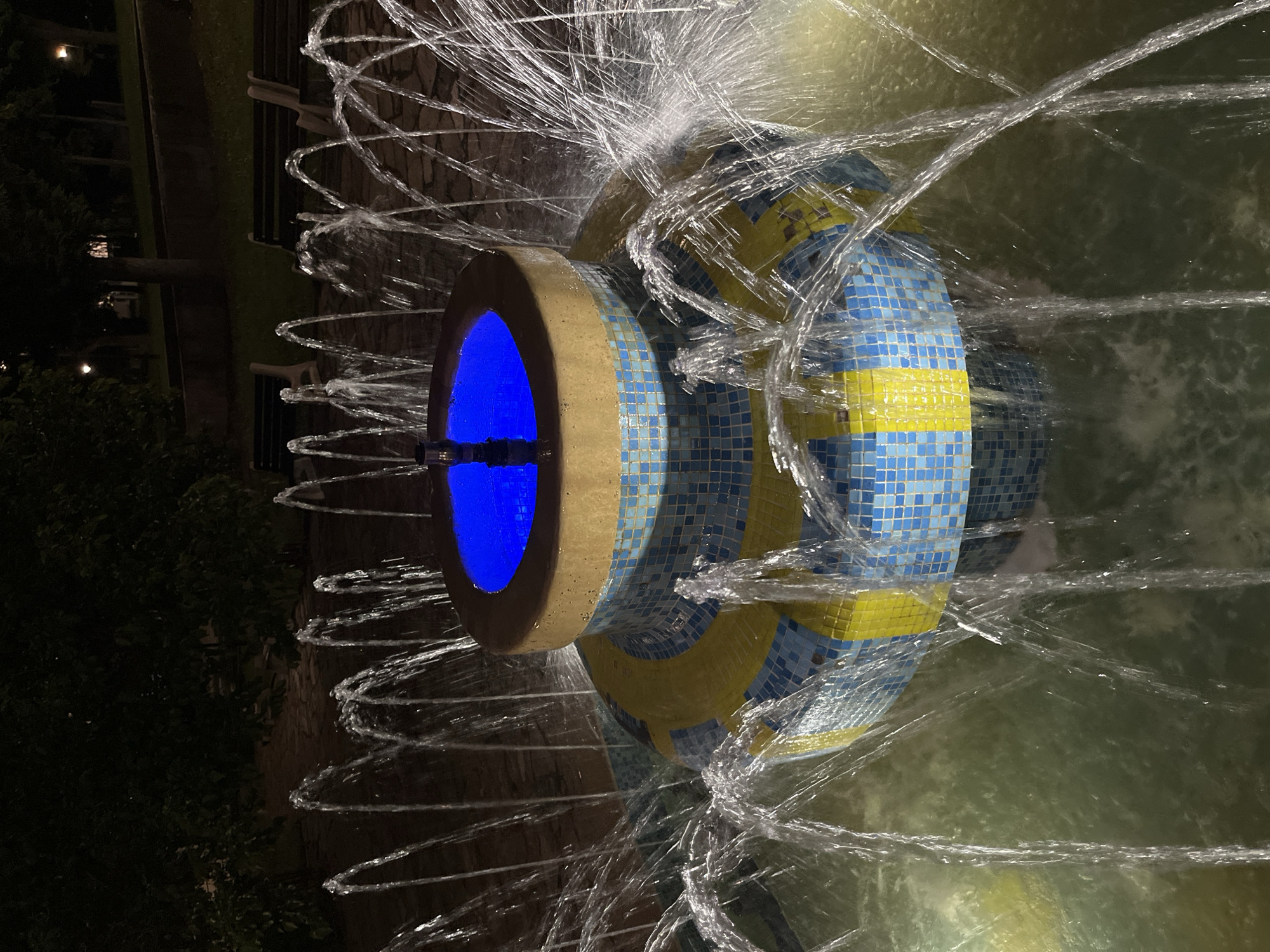 The image displays a multi-tiered fountain at night, located in Veliki Preslav, Bulgaria. The upper tier of the fountain features a circular basin illuminated by a strong blue light, with a central nozzle or pipe visible. This basin rests on a cylindrical base decorated with a mosaic of small blue and yellow square tiles. Numerous jets of water arc upwards and outwards from various points around the fountain's tiers and from its base pool, creating a dynamic display. The water in the pool appears slightly murky or greenish under the ambient lighting. The background is dark, revealing dense tree foliage and several dark park benches arranged on a paved surface, suggesting a park or garden environment. Faint ambient lights are visible in the distant background through the trees. No individuals are present in the scene.
