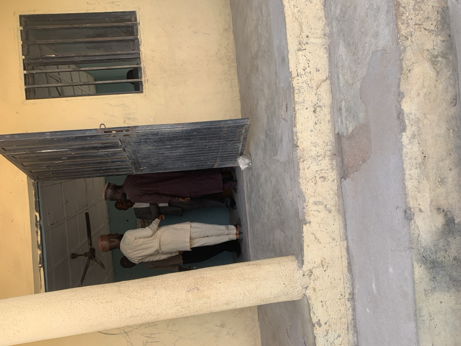 The scene depicts the exterior entrance of a building in Bojude, Nigeria. The building features a worn, light yellow painted facade with a dark metal door, partially open inwards to the right, and a barred window to its right. A large, round, light yellow concrete pillar stands on the left, supporting an overhang. The immediate foreground consists of a two-tiered concrete step structure leading up to a concrete landing at the building's entrance. The concrete surfaces show signs of wear and discoloration. A small, light-colored object, possibly a rock or crumpled paper, rests on the concrete landing near the base of the open door.
Within the open doorway, five adult males are congregated. A man in a light-colored (beige/cream) traditional tunic and trousers, wearing a dark cap, stands with his back largely to the viewer, facing left and slightly inward. To his immediate left, partially obscured by the pillar, is another man in dark clothing. Behind the man in light attire, another man is visible wearing a dark suit jacket. To the right, also facing inward towards the group, is a man dressed in a dark brown or maroon traditional garment and a dark cap. A fifth man in dark clothing is partially visible further inside the doorway, obscured by shadows. Visible inside the building through the doorway is a white ceiling with a dark ceiling fan mounted centrally. The men appear to be engaged in conversation or an informal gathering.