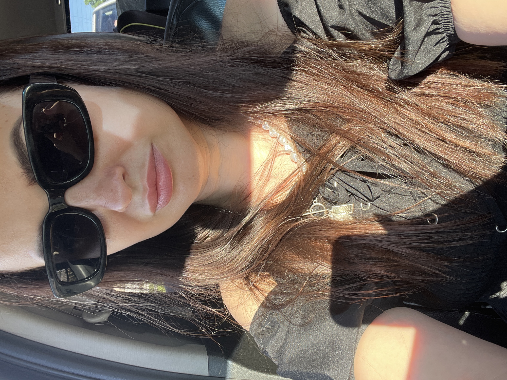 The image displays a close-up portrait of a young woman reclining or lying down inside a car. Her head is positioned towards the upper left of the frame, with her body extending diagonally across the image. She has long, dark brown hair with visible reddish-brown or golden highlights from the sunlight, cascading over her shoulders and torso, which are covered by black clothing, possibly a top or dress with some texture.
The woman is wearing stylish black rectangular sunglasses that cover most of her left eye and part of her cheek. Her lips are slightly parted, hinting at a relaxed expression or a soft smile. Around her neck, she wears a white pearl choker necklace and a longer silver chain necklace featuring a small, flat silver charm. The inscription on the charm is not clearly legible due to lighting and image resolution.
Bright, direct sunlight streams into the car from the left, creating prominent highlights on her hair, skin, and necklaces, and casting distinct shadows on her clothing and the car's interior. The setting appears to be a car's interior, with glimpses of black upholstery and car door trim. Through a window in the upper background, hints of a clear sky and distant trees suggest it is daytime. The woman appears to be posing casually for a photograph.
