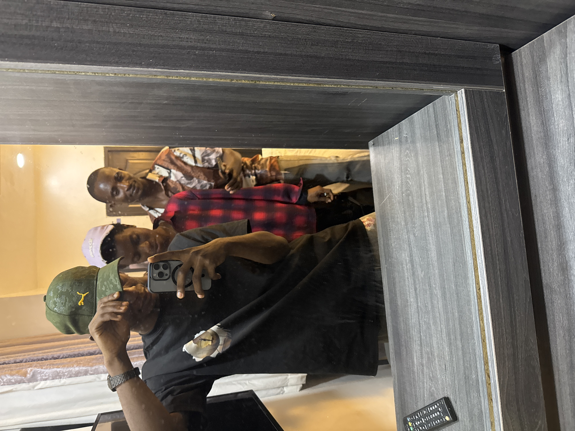 The image shows a reflection in a mirror. The mirror is framed with gray wood. Three men are visible in the reflection. One man is holding a smartphone, presumably taking the photo. He is wearing a black shirt and a green cap. Another man is wearing a plaid shirt, and a third man is wearing a patterned shirt. The setting appears to be indoors. Based on the user's request, the location is Awka, Nigeria, but no evidence in the image confirms this location. A remote control is visible on the right side of the image. The environment is lit artificially. Bedding is visible in the reflection.