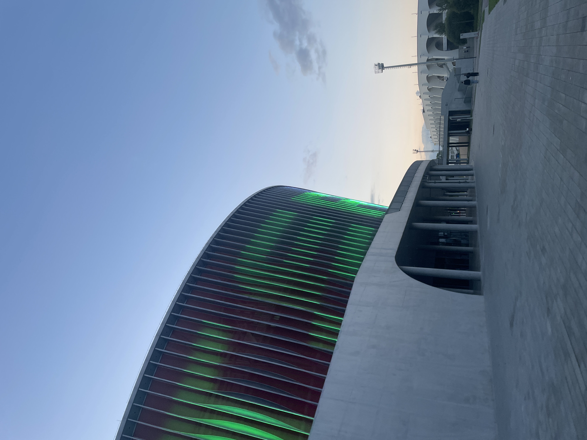 The image shows a modern stadium with a curved facade illuminated by green lights. The stadium has a striking architectural design with horizontal metallic louvers on the exterior. Adjacent to the stadium is a paved walkway made of interlocking gray bricks. In the background, another part of the stadium complex is visible, featuring a white, multi-tiered structure with arched openings, possibly seating areas or support facilities. A tall lighting tower stands to the right of this structure. The sky is a clear blue with a few wispy clouds towards the horizon, suggesting it is either early morning or late afternoon. The lighting on the stadium facade indicates that it is illuminated for an evening event or display. There are two figures walking on the paved area in the distance, appearing small against the scale of the stadium. The overall impression is one of contemporary architecture and an urban setting. There is no visible text.