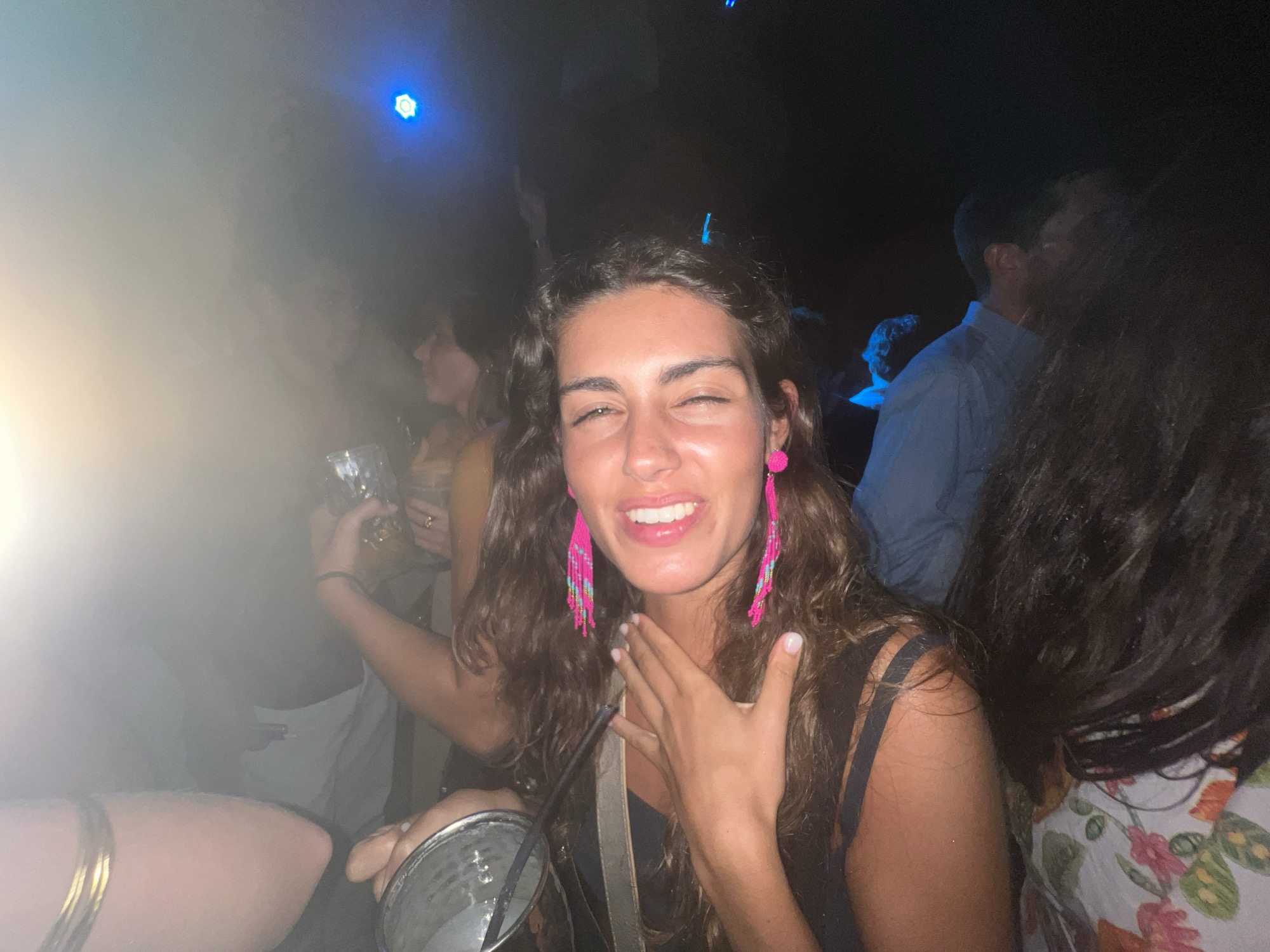 This image depicts a lively outdoor gathering at night, possibly a party or festival, in Quarteira, Portugal. The foreground is dominated by a young woman with long, wavy brown hair and prominent pink, beaded earrings. She is smiling broadly and looking towards the camera, with one hand raised to her chest. She is wearing a dark top. To her left, another person, partially visible, holds a drink. In the background, the scene is illuminated by blue spotlights and other lights, creating a hazy, atmospheric effect. Several other individuals are present, their forms blurred by the low light and movement. The overall impression is one of a dynamic and joyful social event.