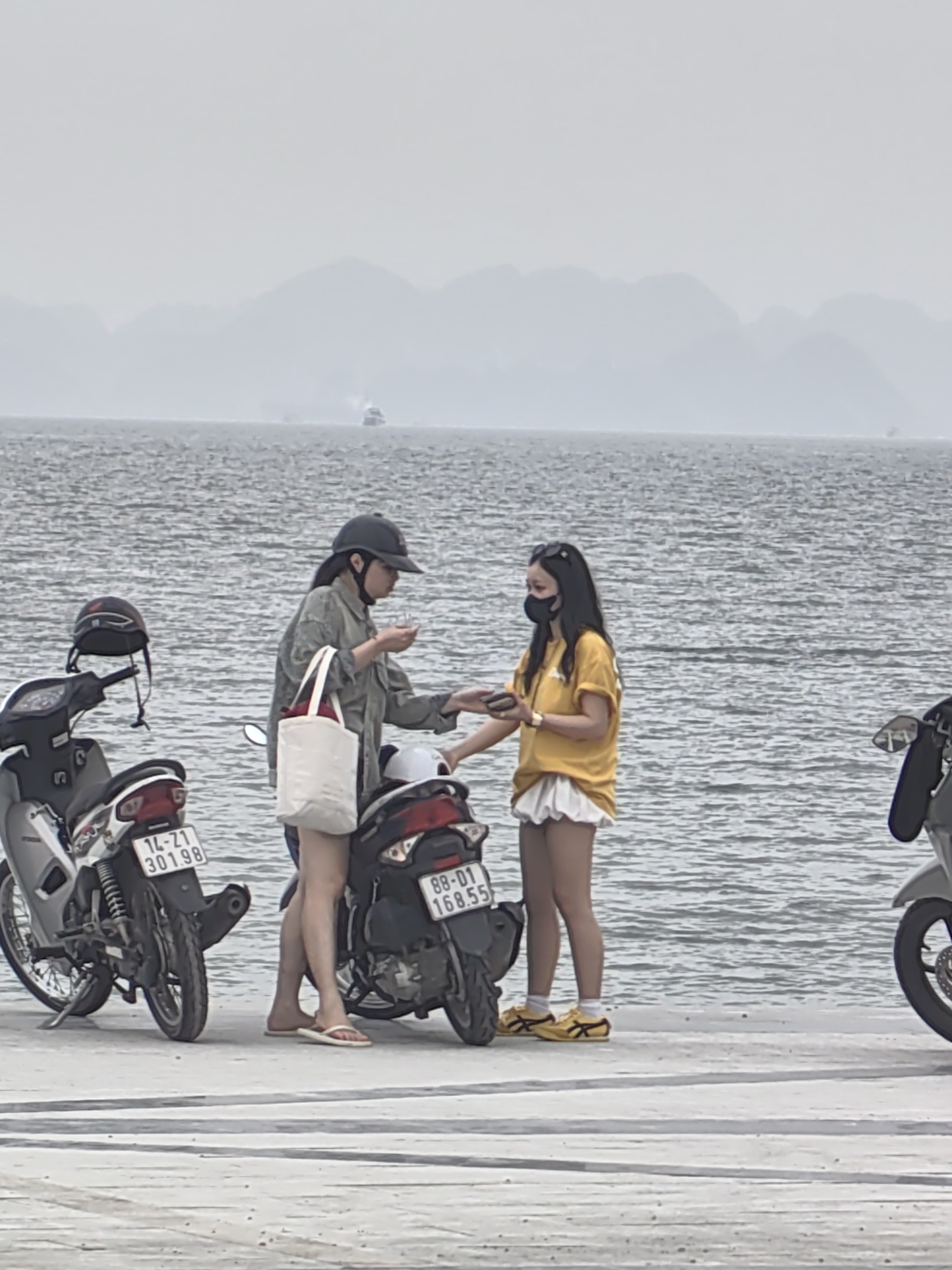 Two young women are standing on a concrete pier in Bai Chay Ward, Vietnam, interacting near two parked motorbikes, with the ocean and distant mountains in the background. The woman on the left wears a grey jacket, light shorts, flip-flops, and a black baseball cap, holding a white tote bag; she appears to be passing an item to the other woman. The woman on the right wears a yellow shirt, white shorts, yellow shoes, sunglasses on her head, and a black face mask. A grey motorbike with a helmet rests on the pier next to the women. A second motorbike is parked nearby. The license plates are visible on the bikes. The sky is overcast and the ocean stretches to the horizon with a few islands or mountains visible in the distance.