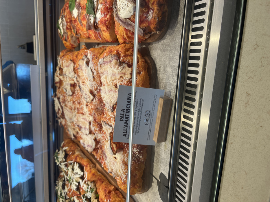 The image displays a close-up of various Italian pizzas, specifically "pizza alla pala" (pizza by the shovel), showcased in a refrigerated or heated display case within what appears to be a food establishment in Dorno, Italy.
The main subject is a large rectangular pizza labeled "PALA ALL'AMATRICIANA." This pizza features a thick, golden crust and is generously topped with a red sauce, chunks of what is identified as guanciale (cured pork cheek), and a generous dusting of grated Pecorino Romano DOP cheese and black pepper.
Above the Amatriciana pizza, there are other types of pizza. One visible portion features anchovies and fresh green basil leaves on a red sauce base, while another has anchovies on a creamy white base, possibly with a dollop of soft cheese. Below the Amatriciana pizza, a section of another pizza is visible, topped with green herbs and white grated cheese.
A white price tag with a wooden base stands in front of the "PALA ALL'AMATRICIANA." The visible text on the tag reads:
- "PALA ALL'AMATRICIANA"
- "Pizza"
- "pecorino romano DOP e pepe nero"
- "Pizza alla pala with tomato pulp, guanciale, PDO pecorino romano and black pepper"
- "€ 8,20"
Below the price, there is smaller, illegible text, likely detailing ingredients or allergy information. The overall scene suggests a deli or bakery counter where fresh pizzas are prepared and sold.