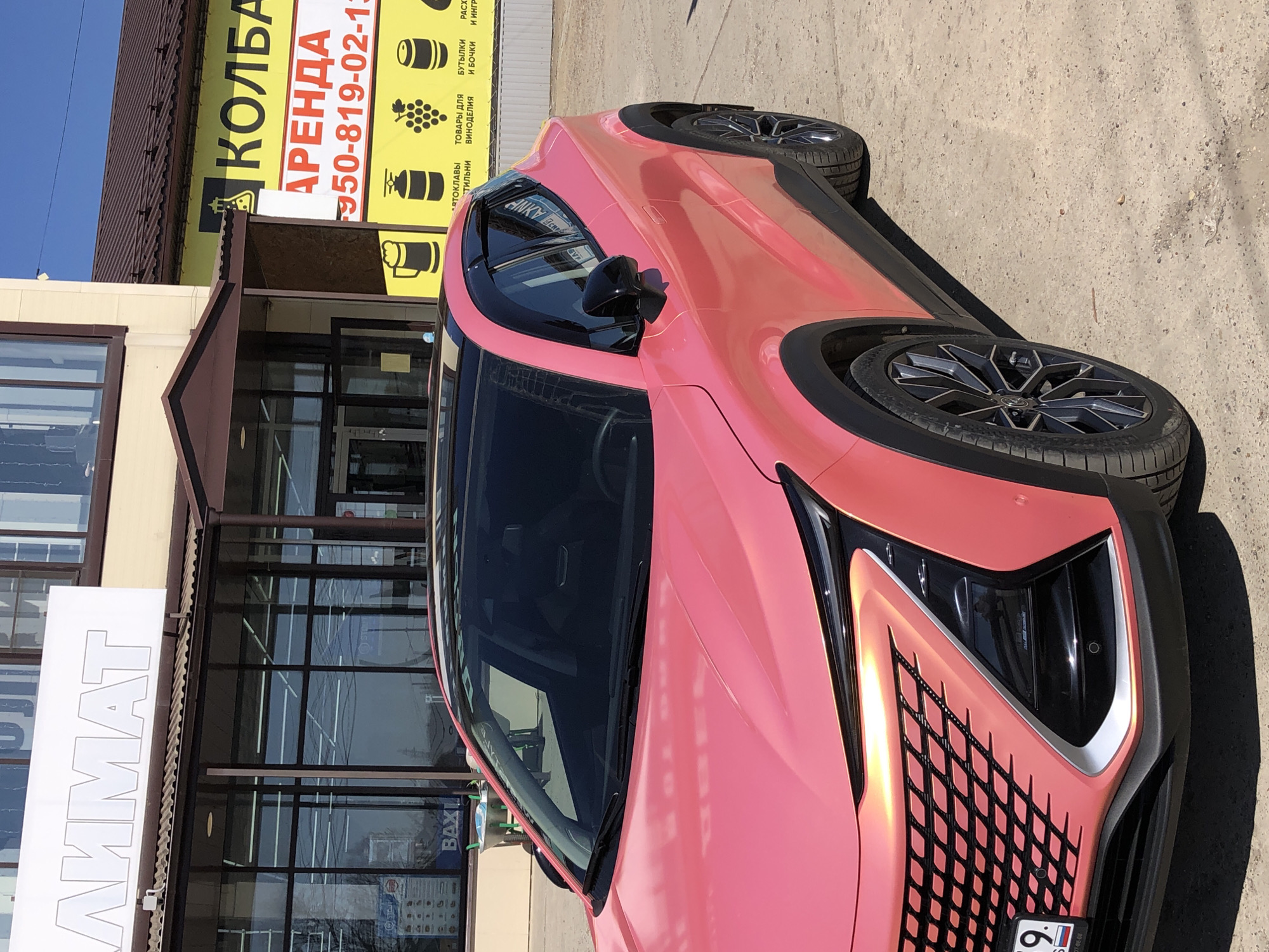 Here's a forensic description of the image:
**General Description:**
The image is a mid-angle shot of a light pink car parked in front of a commercial building. The shot seems to be taken at ground level. The photograph is slightly rotated counter-clockwise.
**Location:**
The building has a sign indicating this is in the city of Chaikovsky, Russia ("АММАТ" can be seen). A separate sign advertises the renting out of space.
**Subjects and Objects:**
* **Car:** The primary subject is a car with a unique light pink color (possibly a wrap). It has dark wheels and a distinct headlight design. The license plate is partially visible.
* **Building:** The building is a commercial structure with multiple glass doors and windows. Signs are visible on the building, including one in Russian advertising "КОЛБА".
* **Signs:** Several signs are visible advertising rental space ("АРЕНДА").
**Environment:**
The scene is outdoors during the daytime with bright sunlight. The ground appears to be a paved surface.
**Overall Impression:**
The image shows a car parked in front of a commercial building, likely a retail space. The area seems to be well-lit and in a commercial or urban setting.