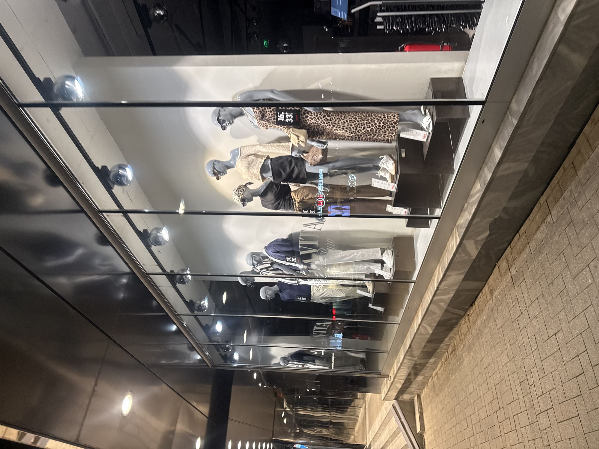 The image depicts a storefront window display in what appears to be an urban environment. Five mannequins are arranged horizontally across multiple tiers within the window. The mannequins are dressed in contemporary casual clothing, with varying outfits including dresses, tops, bottoms, and shoes. The display is illuminated by multiple small, round, silver lights, some of which are reflected in the glass. A reflection of the outside sidewalk and building exterior is visible in the window. There are visible brick pavers of the sidewalk. The store's environment is not clearly indicated but I do not have enough information to provide the location in Varna, Bulgaria.