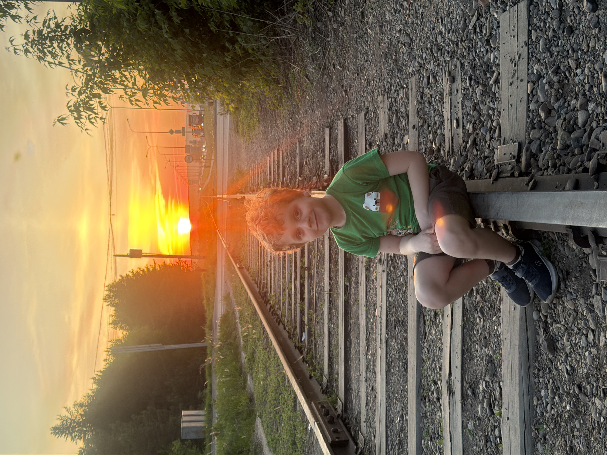 A photograph shows a child sitting on a railroad track at sunset. The railroad tracks extend into the distance with the setting sun directly in line with the tracks. The child, wearing a green shirt and shorts, sits with their legs pulled up to their chest. The tracks are surrounded by gravel and sparse vegetation. Beyond the tracks is a road with street lights and trees. The setting appears to be Chaikovsky, Russia, given the style of street lighting and the vegetation.
