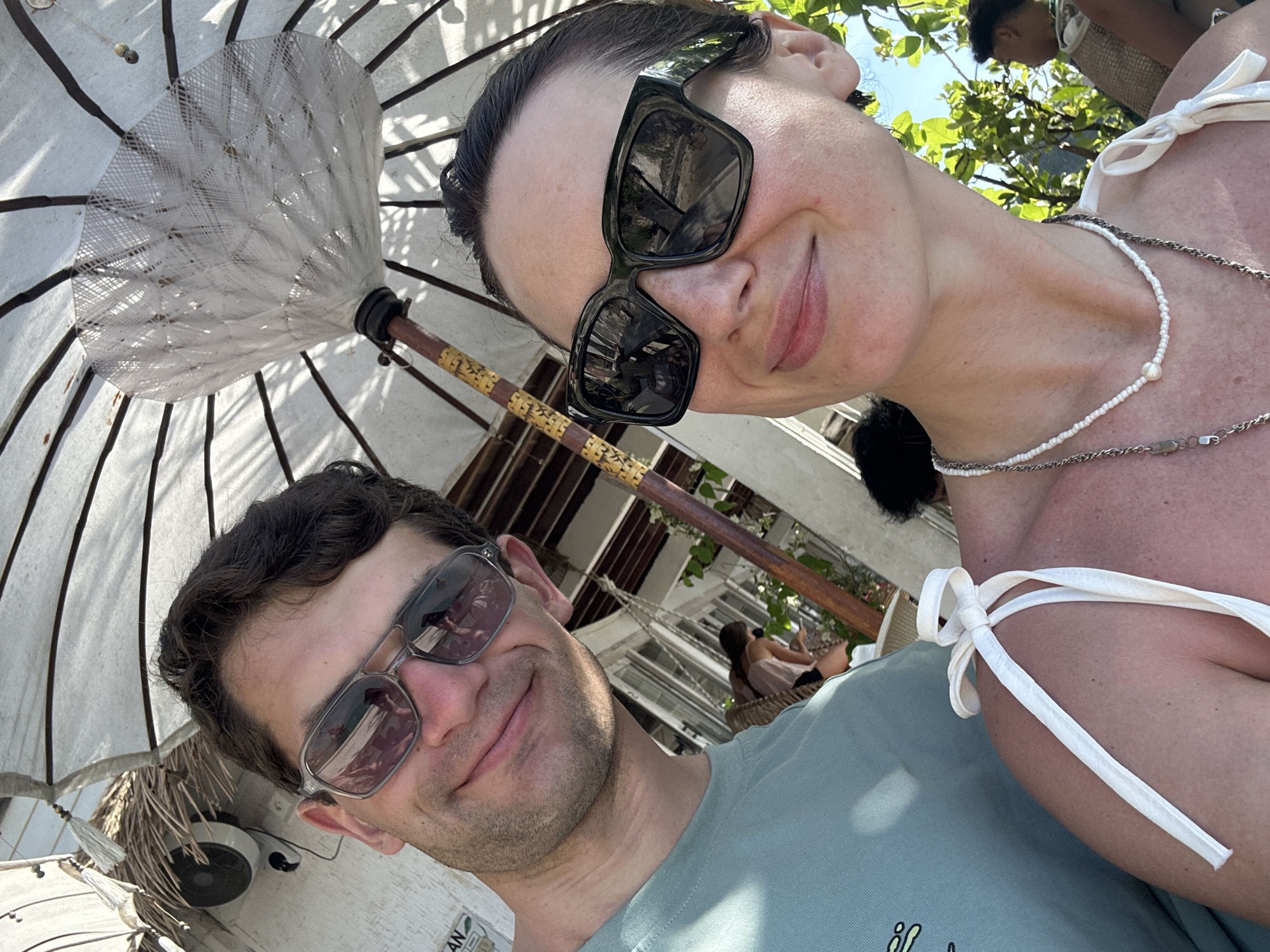 A selfie shows two individuals under a white umbrella with a light wooden pole. The location is Kuta Selatan, Indonesia, based on an inscription on a white sign at the bottom left of the frame. The man, wearing a light green t-shirt and sunglasses, is positioned on the left. The woman, wearing large black sunglasses and a white bikini top, is on the right. She also wears two necklaces. In the background are building details and foliage. Another woman sits in a chair in the background.