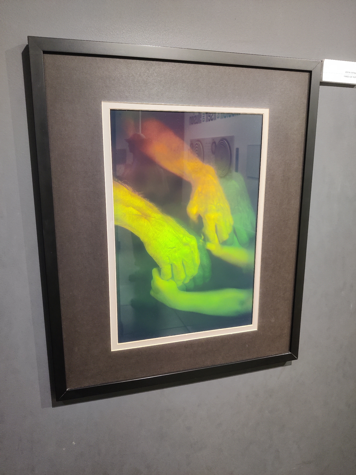 The image shows a framed artwork hanging on a grey wall in Thessaloniki, Greece. The artwork is a photograph with a prominent display of hands, rendered in vibrant yellow and green hues, creating an abstract and somewhat ethereal effect. Two sets of hands are intertwined, with one pair appearing to be helping or holding the other. The background of the photograph is dark and blurred, with some faint reflections of what appear to be circular shapes. A small white label is attached to the wall to the right of the frame, with Greek text and the English translation "HANDS OF HOPE." The lighting in the scene is ambient, with some light reflecting off the glass of the frame. The overall impression is one of a contemporary art exhibition.