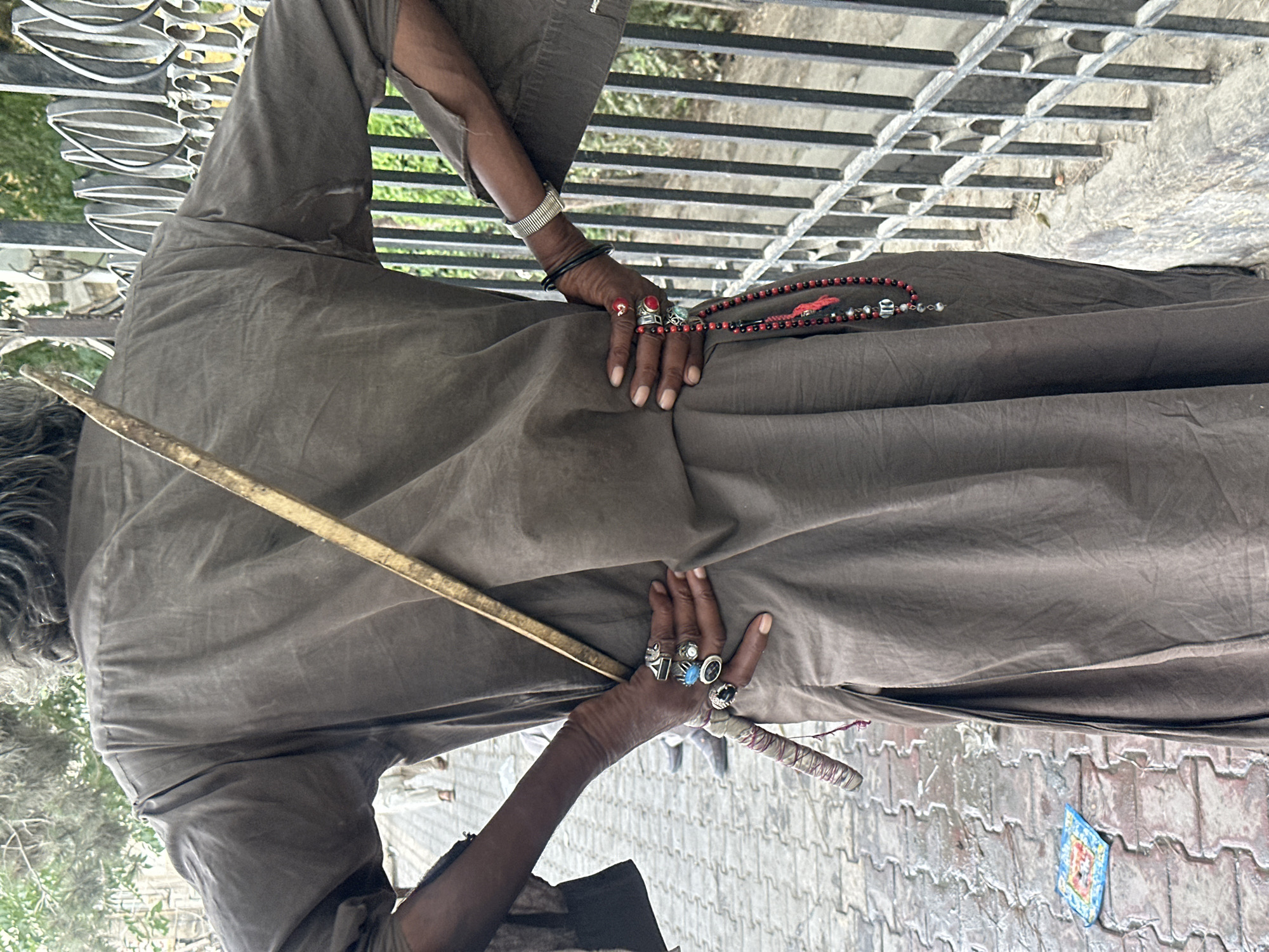 The image depicts the back of a person wearing a loose, dark-colored garment, possibly a chugha or kurti, with a wooden stick resting diagonally across their back. The person has several rings on their fingers, including large statement rings with stones and ornate metalwork. A strand of dark beads with red accents hangs from their right hand. Their left arm is bent, with their hand resting on their hip. On their right wrist, they wear a metallic cuff bracelet. The setting appears to be an outdoor urban environment. In the background, a black metal fence with decorative elements is visible, along with some greenery. The ground is paved with interlocking bricks, and there is a small, colorful object on the ground near the lower left corner. The overall impression is of a person in a street setting, possibly in Quetta City Tehsil, Pakistan, given the style of dress and adornments.