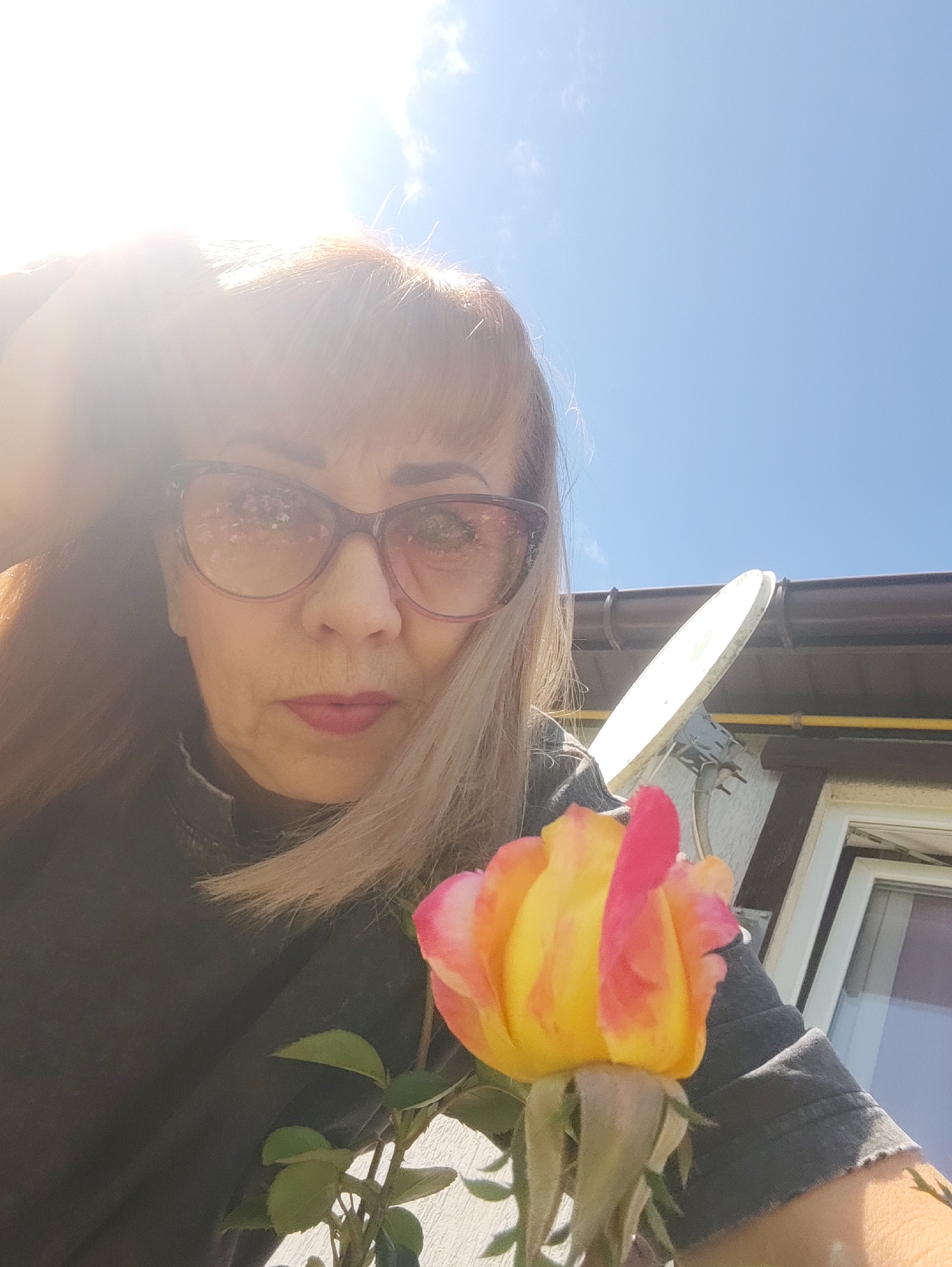 The image is a selfie taken outdoors, possibly in a residential area, under a clear blue sky. A woman with blonde hair, wearing large, reddish-brown sunglasses and a dark grey top, is holding a yellow and pink rose in the foreground. The rose is partially in focus and close to the camera, with green leaves visible. In the background, there is a house with brown trim and a white satellite dish mounted on the wall. The sun is bright and positioned in the upper left corner, creating a lens flare effect. There are no specific landmarks visible that would definitively place the image in Gvardeysk, Russia.