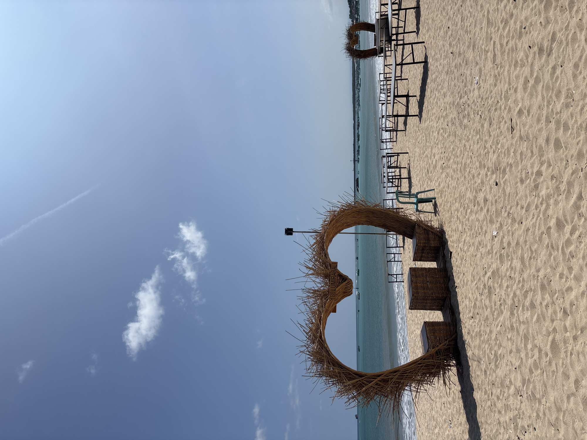 The image shows a beach scene in Kuta, Indonesia, under a clear blue sky with a few scattered clouds. The foreground features a large heart-shaped structure made of natural materials like straw or reeds, with a small sign attached to its upper portion. Wicker seating is visible beneath the heart. The beach extends into the distance with numerous tables and chairs set up, suggesting a dining or lounge area by the sea. The ocean is calm, with gentle waves lapping the shore. The overall impression is a serene beach setting with a romantic or decorative installation.