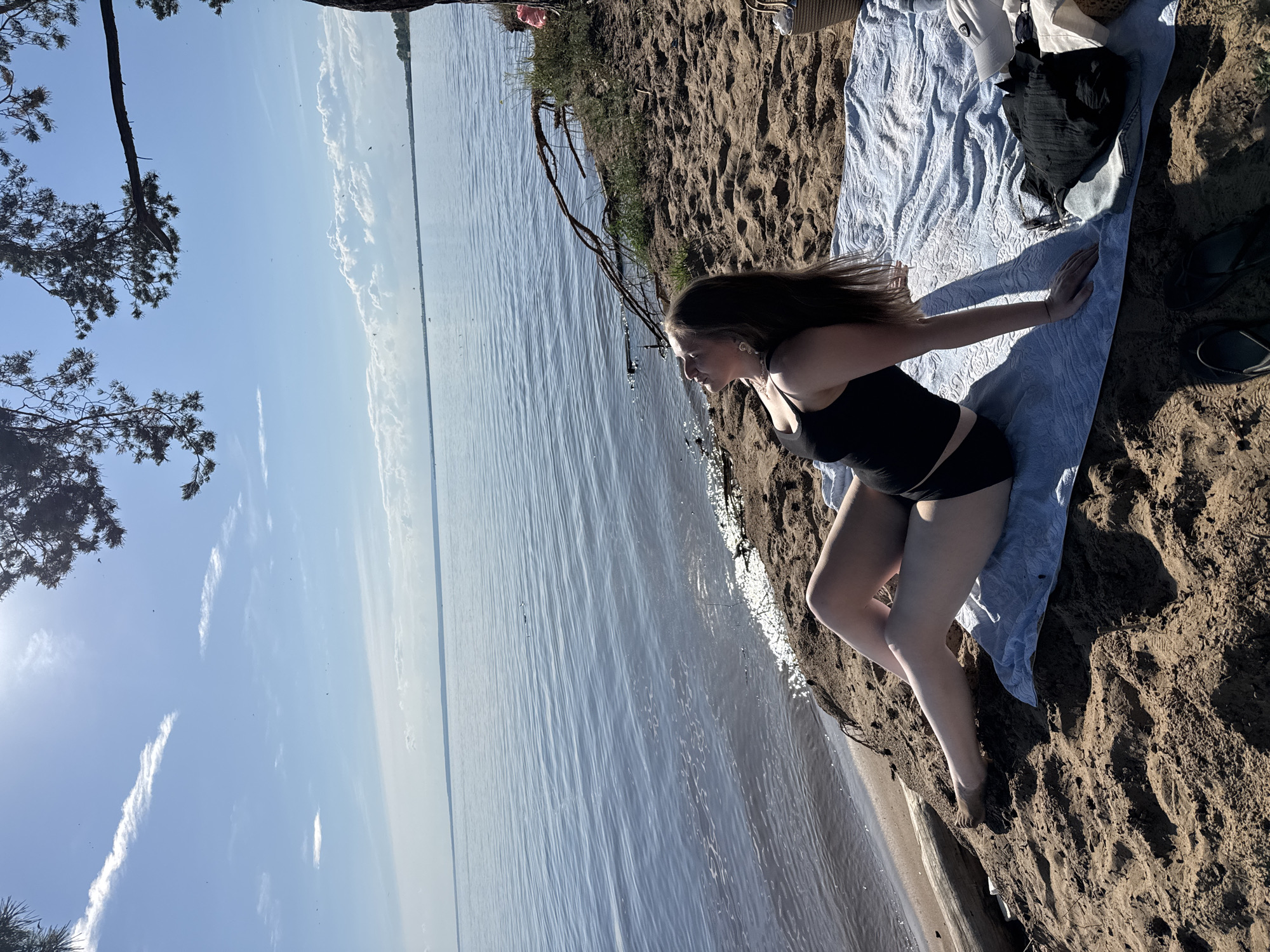 A woman is sitting on a beach on a towel, facing the water and sky. She is wearing a black swimsuit. The beach is sandy with some driftwood. The water is calm and blue, with a clear sky overhead. There are some trees with branches extending over the scene. There are also some clothes, a hat, and a bag next to the woman.