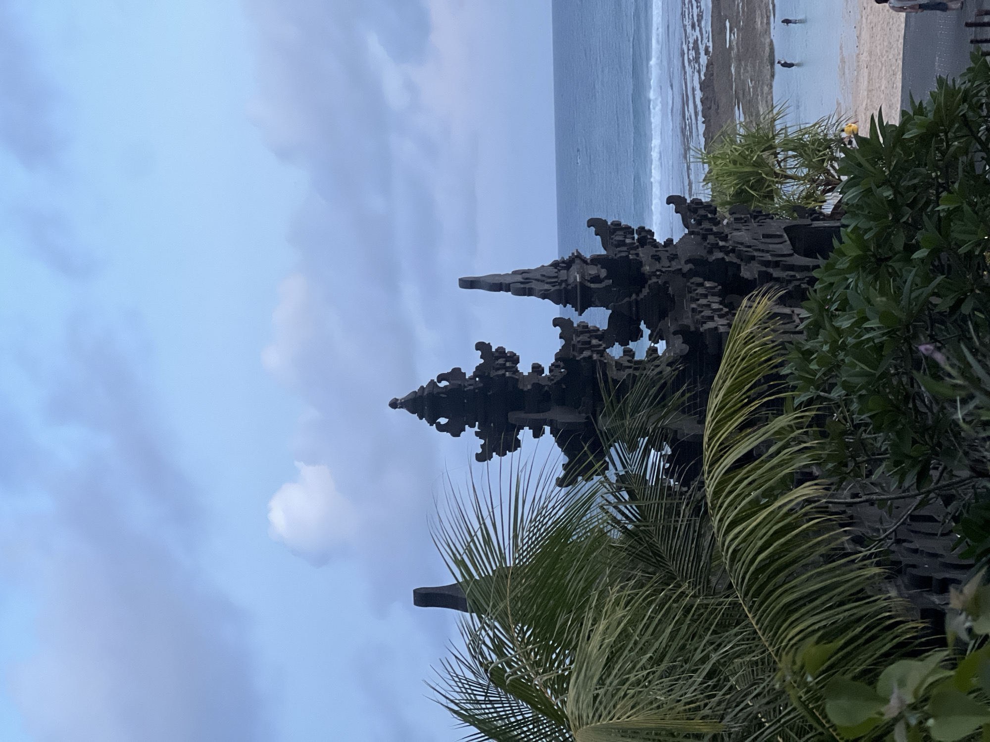 Captured from a high-angle, tilted perspective, the image presents a coastal scene, likely in Kuta Selatan, Indonesia. The foreground features lush tropical foliage, including palm fronds. Behind the vegetation, a dark stone structure rises, its architectural details suggest it may be part of a temple or religious site. In the midground, the ocean stretches out, with waves breaking near the shore. The shoreline is rocky in places, transitioning to a sandy beach. A few individuals are visible on the beach, some standing in the water. The sky is overcast, with clouds filling much of the frame.