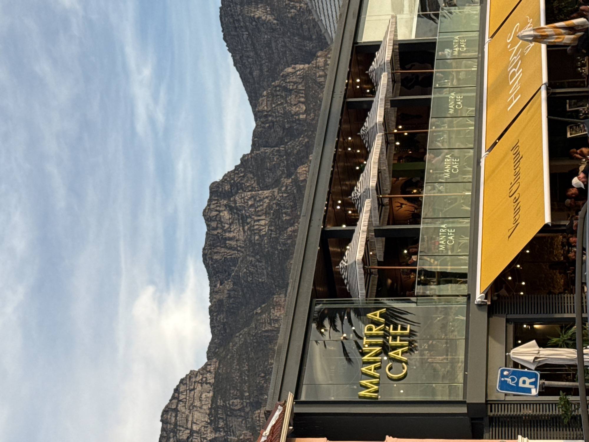 The image shows a partial view of Mantra Cafe in Cape Town, South Africa, set against the backdrop of a mountain. The cafe is a modern, multi-story building with glass walls bearing the cafe's name in gold lettering. Each level of the cafe has awnings. The interior of the cafe is visible through the glass, revealing patrons dining. To the right of the cafe entrance is a yellow awning with the words "Henry's" on it. A handicapped parking sign is also visible. The sky is blue with streaks of clouds. The mountain is rugged and textured.