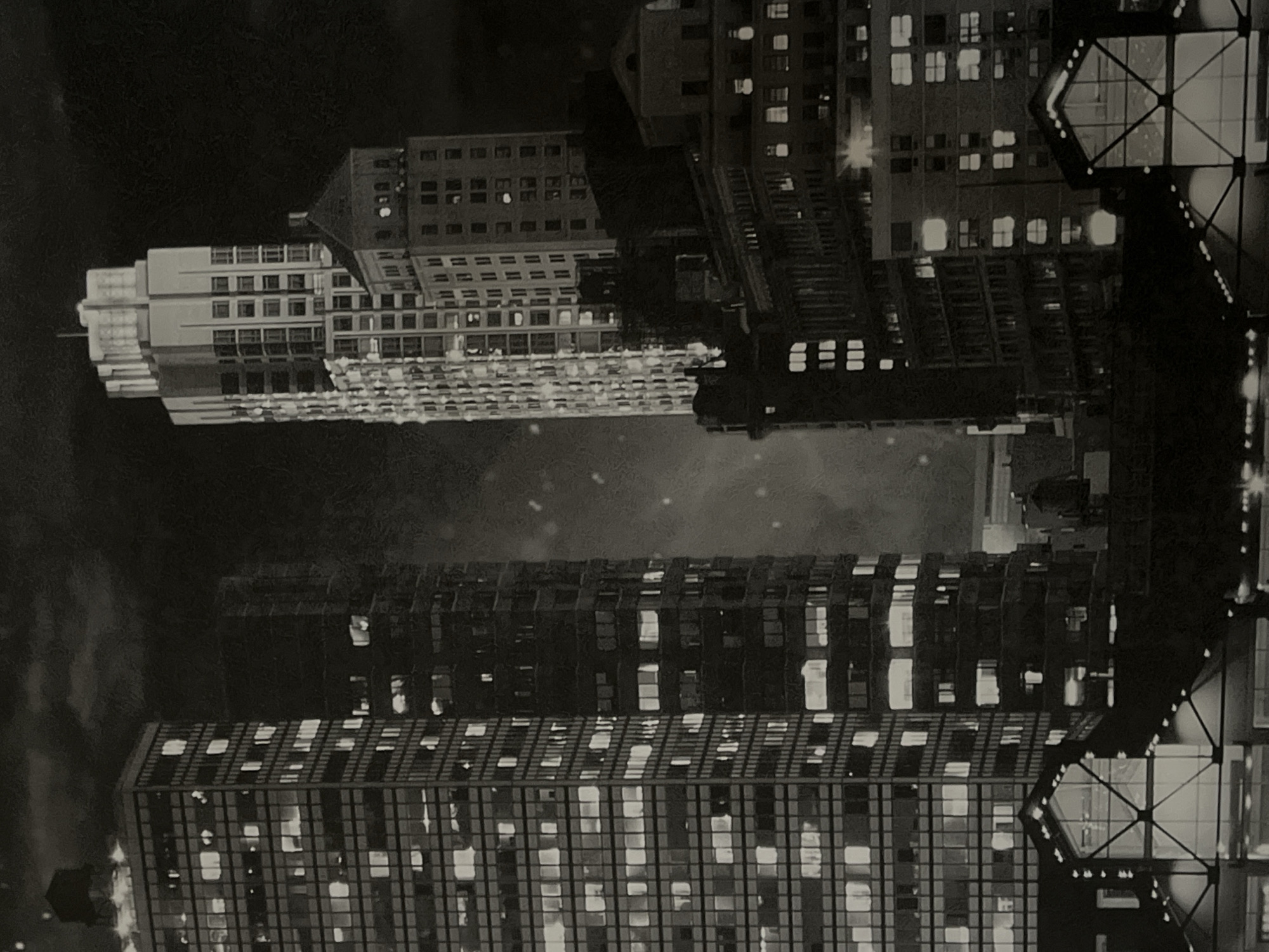 This is a black and white, high-angle, nighttime photograph of a cityscape featuring multiple skyscrapers. The buildings are illuminated by numerous lit windows, suggesting an urban environment with active businesses or residences. The sky is dark with subtle clouds, and there are some indistinct bright spots that could be stars or distant lights. In the foreground, there are silhouetted structures with geometric designs, possibly rooftops or architectural elements of lower buildings. The overall impression is one of a bustling metropolis at night. The location is identified as the city of Dnipro, Ukraine.