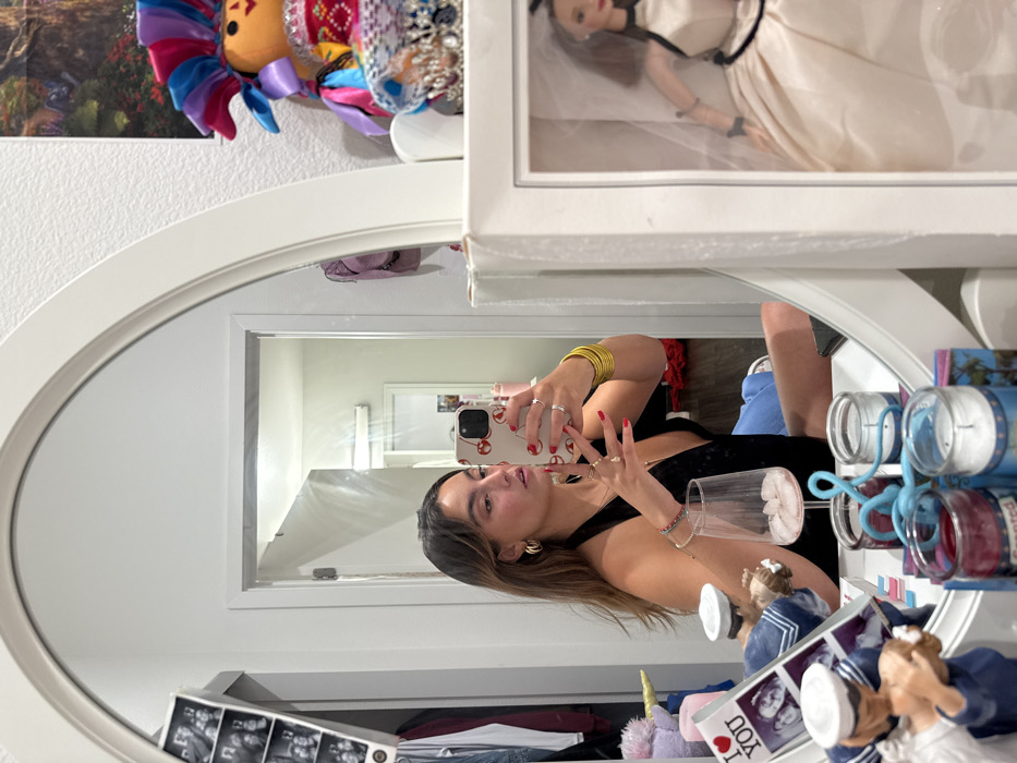 A woman with brown hair and gold hoop earrings is taking a selfie in an oval mirror. She is wearing a black tank top, gold bracelets, and blue jeans. Her phone has a red and white design. The mirror reflects a hallway with a white door. Around the mirror are decorative items, including a doll in a wedding dress, a colorful toy, and a photo album. The overall scene appears to be a cluttered but stylish bedroom.