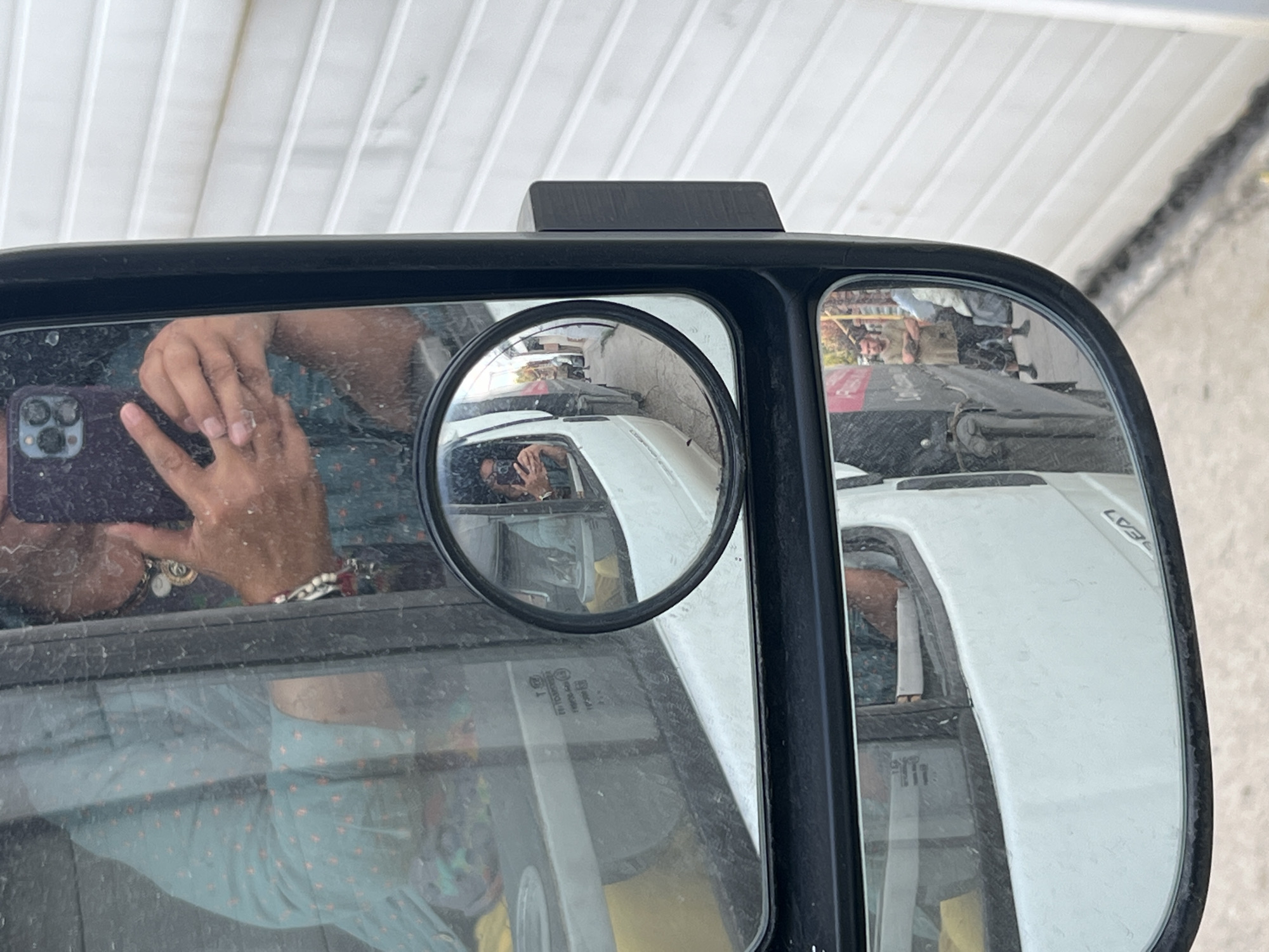 The image is a close-up, reflection-heavy shot from the perspective of a person inside a vehicle, looking out of the side mirror. The primary focus is the side mirror of what appears to be a white van, revealing reflections of the surrounding environment. In the larger section of the mirror, a person is visible, holding a smartphone to take a selfie. This person is wearing a patterned blue and green shirt and several necklaces. The reflection also shows the interior of the van, with some yellow material visible.
A smaller, circular inset mirror shows a more distant view. In this reflection, the same person taking the selfie is visible in the van window, and further down the street, two men are standing outside. One man, wearing a tan t-shirt and dark shorts, has his arms crossed and is looking towards the camera. The other man is partially obscured. The background in the inset mirror consists of a white, corrugated building and a street or alleyway.
The overall scene suggests a street setting, likely in the city of Ozernyy, Russia, given the prompt. The reflections create a layered and somewhat disorienting view, with the observer's presence implied by the selfie-taking action. The image has a slightly gritty texture, possibly due to dirt or rain on the mirror or windows.