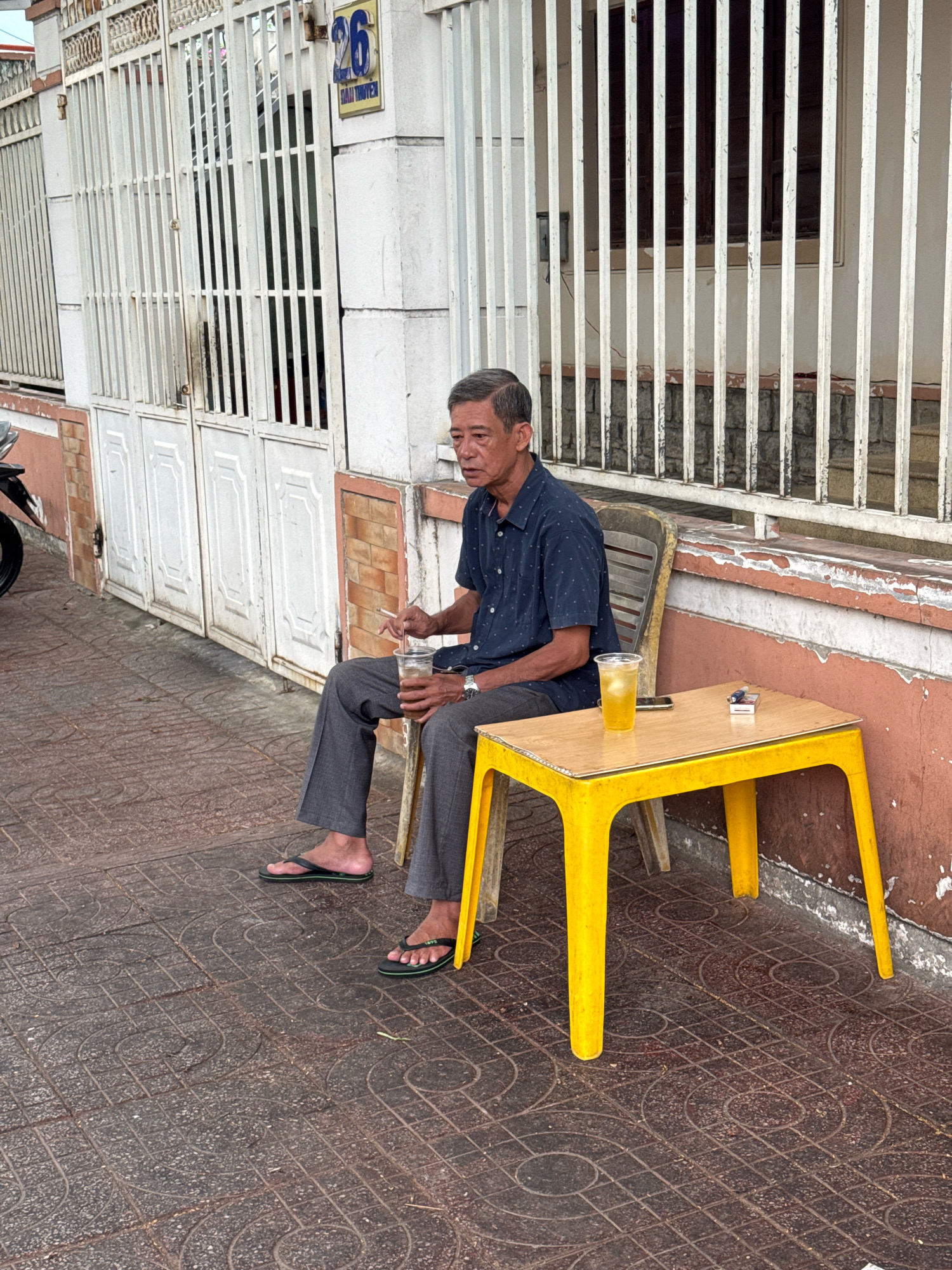 A middle-aged Vietnamese man is seated outdoors on a plastic chair, holding a disposable cup with a straw. He is wearing a dark blue short-sleeved shirt and grey trousers, and black flip-flops. A small yellow plastic table is placed in front of him, on which sits a glass of yellow liquid, a pack of cigarettes, a lighter, and a mobile phone. The man appears to be smoking. The man is sitting outside a building with a white barred gate and a brick-patterned wall. A street number "26" is visible on the building. The ground is paved with patterned tiles. The scene is likely located in Nha Trang, Vietnam.