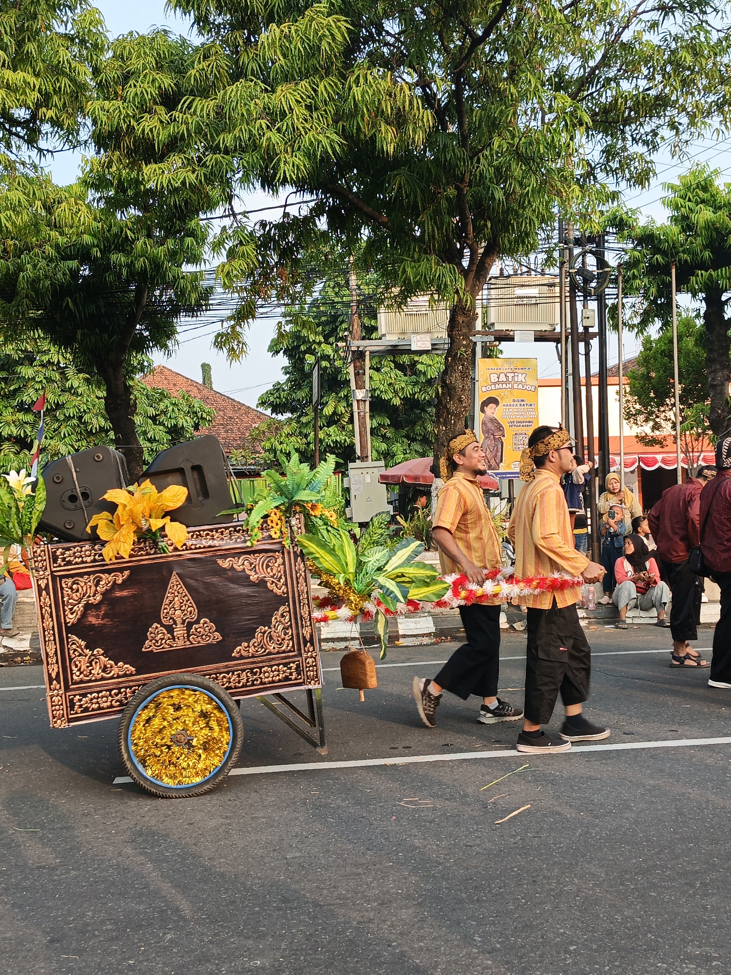 In Klaten, Indonesia, a street scene unfolds with a procession. Two men, adorned in traditional yellow and orange striped shirts with gold headbands, walk side-by-side. They are carrying a decorative garland made of green leaves and white and red flowers, which is suspended between them by thin white ropes. Behind them, a decorative cart is being pushed. The cart, with ornate dark wood paneling and gold detailing, features two large black speakers and a vibrant arrangement of yellow and green artificial flowers. A single wheel of the cart is visible, covered in gold tinsel. Spectators, including women and children, are gathered on the right side of the road, observing the procession. One child is holding a phone, likely taking a photo. Buildings with orange-tiled roofs and lush green trees line the background. A large banner promoting "Batik Kosmetik" is visible, featuring a woman in traditional attire. The scene appears to be a cultural event or parade taking place on a paved street under a clear sky.