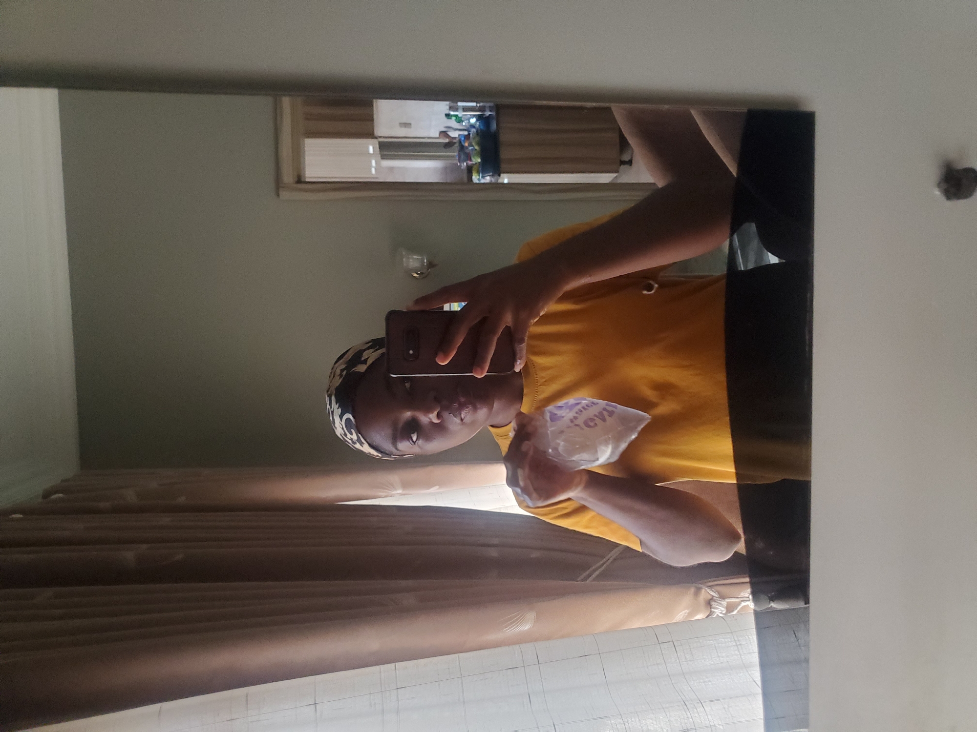 This image, located in Barden Yana, Nigeria, depicts a young adult female captured in a mirror selfie. The subject is standing indoors, wearing a mustard yellow short-sleeved t-shirt with a visible drawstring loop on the lower left hem, and a patterned headwrap featuring black and light-colored swirling designs. Her skin tone is dark. Her right hand holds a dark-colored smartphone, positioned vertically, used to take the photograph, with the phone's camera array visible. Her left hand holds a small, clear plastic bag containing a white granular substance, possibly sugar or salt. Mirrored purple text, partially legible as "OVIA" or similar, is visible on the bag.
The background, reflected in the mirror, shows an interior room with light grey/greige walls and a white ceiling. To the left, a window is covered by two layers of curtains: an inner sheer, light-colored curtain with a subtle grid pattern, and an outer opaque, brown/tan curtain with vertical folds. A wall-mounted light fixture, appearing as a silver or chrome sconce, is visible on the wall behind the subject's left shoulder. The bottom edge of the mirror features a dark, reflective horizontal surface, possibly a shelf or frame, below which the light grey wall continues. A small, dark, irregular mark is present on the lower wall beneath the mirror.
To the right, a doorway with a light-colored wooden frame reflects into another room, which appears to be a kitchen or utility area. This reflected room contains dark countertops, light brown wooden cabinets below the counter, and a dark blue/green bowl filled with light green fruits (possibly limes or small apples) resting on the countertop. Various other small containers and bottles are also visible on the countertop and shelves within this reflected space.