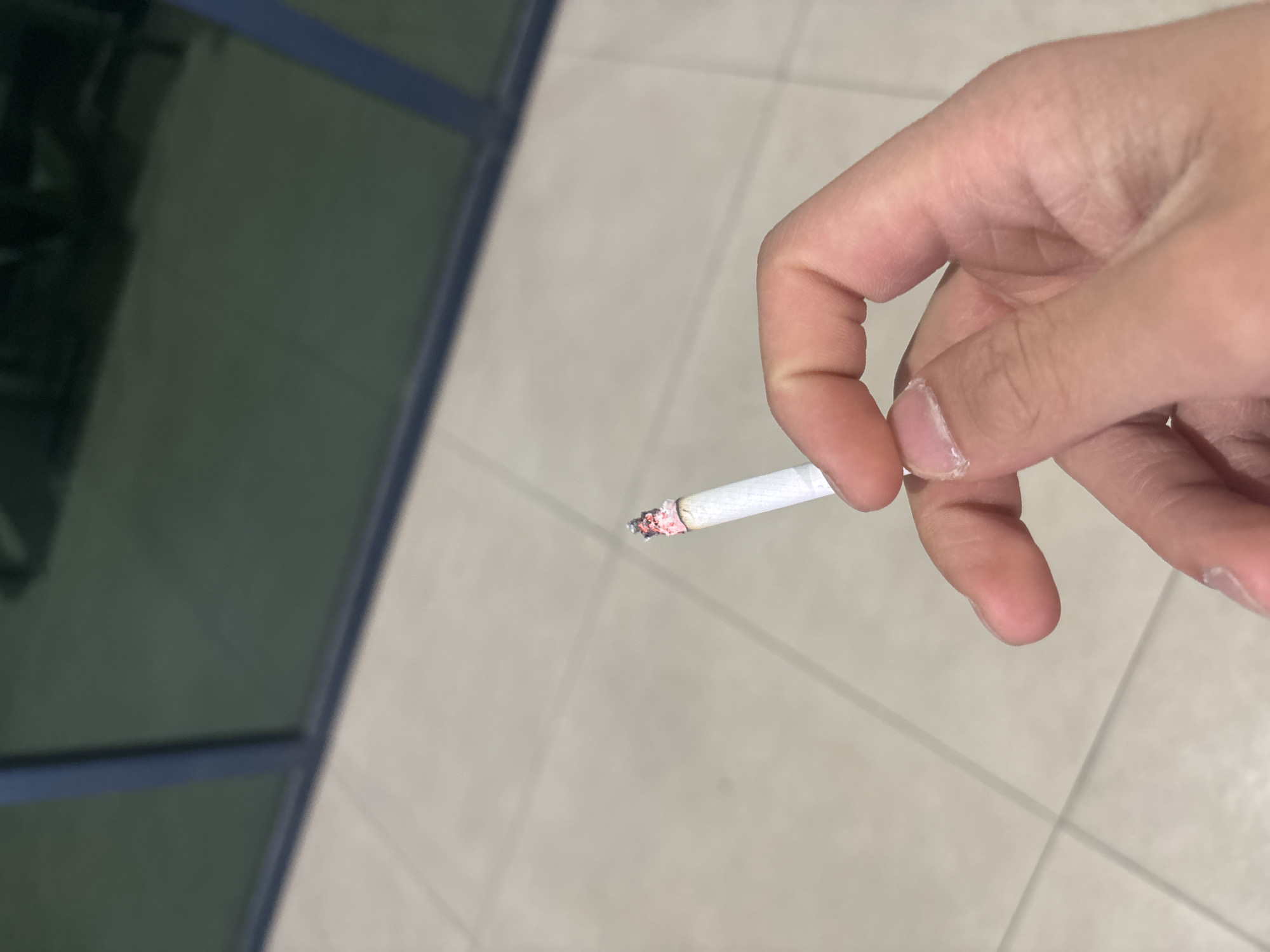 A close-up shot captures a hand holding a lit cigarette, viewed from an overhead perspective. The hand, with fingers gently curled, occupies the right side of the frame, firmly grasping the white cigarette. The tip of the cigarette is visibly glowing with burning embers, and a small amount of ash has formed.
The background is split into two distinct areas. To the left, a dark, reflective surface, likely a window or glass partition, shows a blurred, deep green-grey reflection. A prominent blue line runs across this surface, suggesting a frame or structural element. To the right and below the hand, a light-colored, possibly beige or pale grey, tiled floor is visible, featuring rectangular tiles with discernible grout lines forming a grid pattern.
The overall setting appears to be an indoor or semi-enclosed environment, such as a hallway or a covered area. The dimness of the reflective background suggests it may be evening or night, or an interior space with low natural light. There are no other subjects, activities, or visible text present in the image. The context of Yerevan, Armenia, suggests this scene takes place within that city.