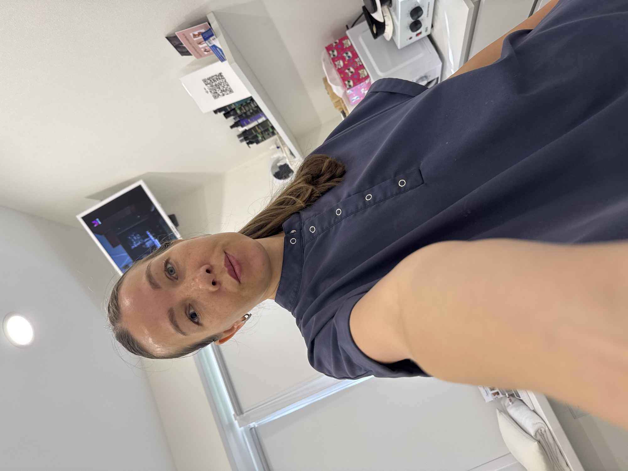 A woman is taking a selfie in a room that appears to be a beauty salon or treatment room. She is wearing a dark blue, collared, short-sleeved shirt with white buttons. Her hair is pulled back into a ponytail. In the background, there are shelves with various bottles, a small television screen on the wall, a white machine with knobs, and folded white towels on a shelf. The room is well-lit and appears clean and organized. The image does not provide any information about the city of Chaikovsky, Russia.