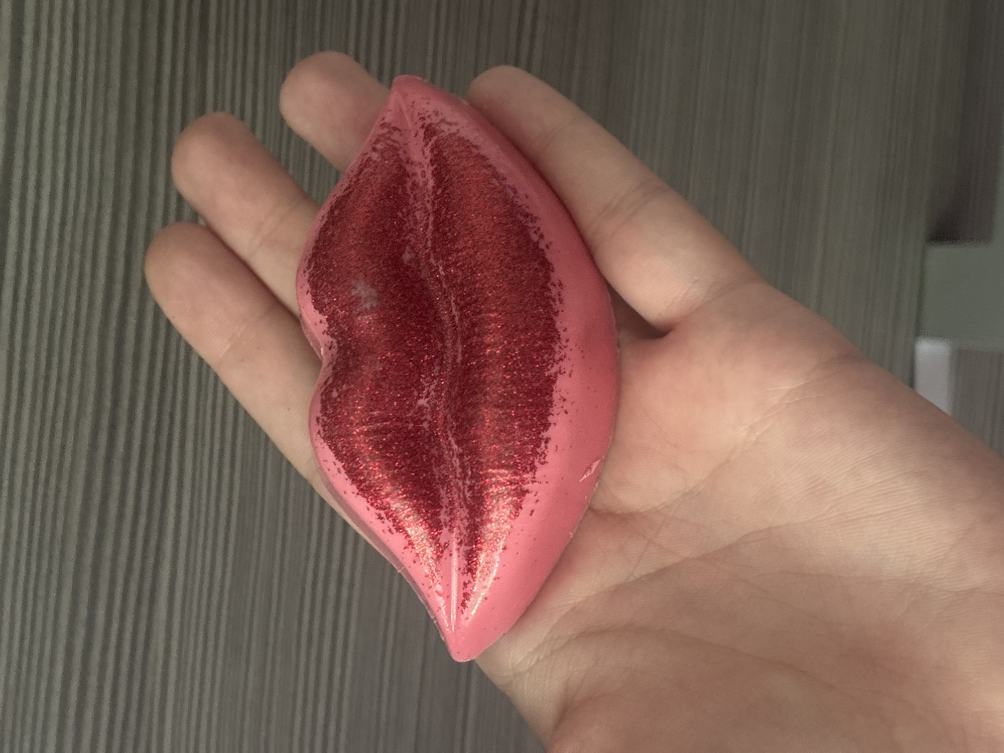A hand holds a pink, glittery object shaped like a pair of lips. The background is a gray, horizontally-lined surface.