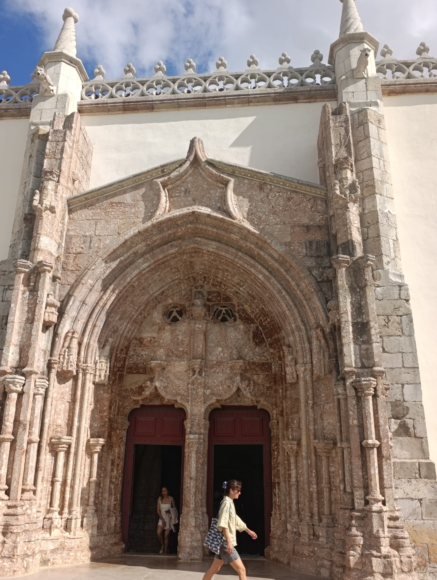 The image captures a sunny day with a bright blue sky dotted with white clouds, framing a section of a grand stone building, likely a church or historical monument, in Setúbal, Portugal. The focus is on an ornate, weathered stone archway, intricately carved with decorative elements and framed by fluted columns. The stone exhibits a reddish-brown hue with patches of lighter and darker discoloration, indicative of age and exposure.
Within the archway are two large, dark red wooden doors. The left door is slightly ajar, revealing a glimpse of the dark interior and a woman in a light-colored dress standing just inside. She has dark hair and appears to be looking down.
To the right of the archway, a woman with light-colored clothing and dark hair is walking away from the entrance. She is wearing sunglasses and carrying a patterned bag. Her stance suggests she is moving casually. The architectural style is elaborate, with Gothic-inspired tracery visible along the upper edge of the building and a pointed finial atop a spire on the right. The stonework itself shows signs of erosion and texture, adding to the historical feel of the structure. The ground in front of the entrance appears to be paved.