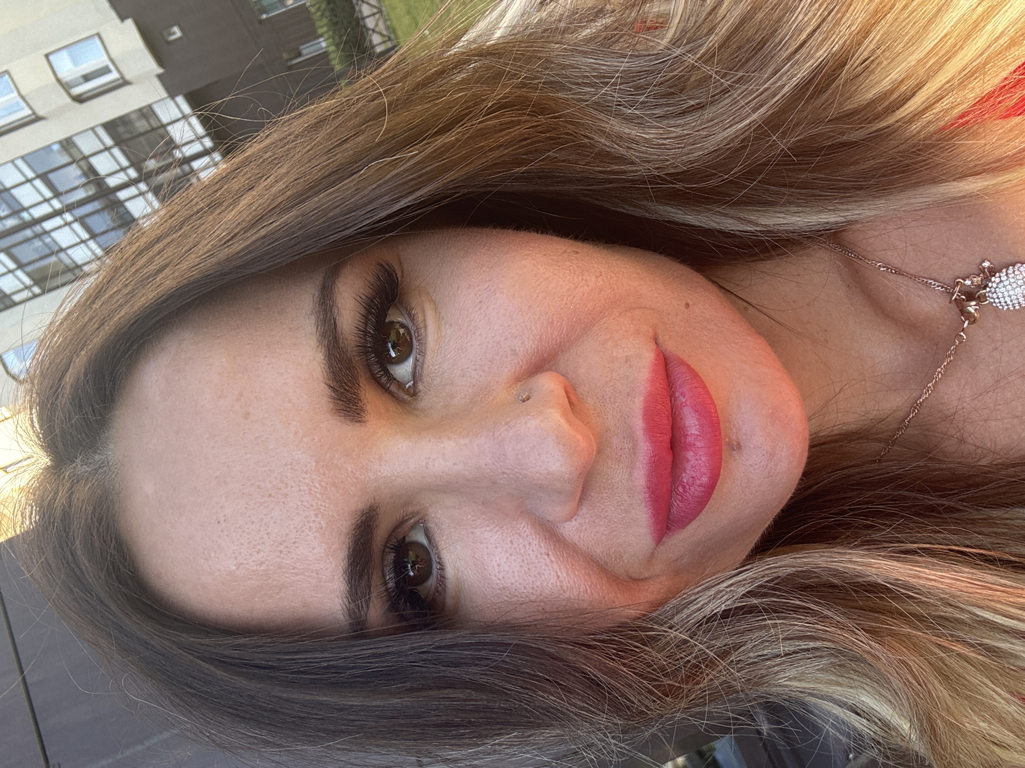 This is a close-up, vertically oriented photograph of a woman's face, tilted on its side. The woman has long, wavy hair that transitions from dark brown to lighter blonde tones, and she is wearing makeup, including bright pink lipstick and false eyelashes. A delicate rose gold necklace with a diamond-encrusted pendant is visible on her neck. A small nose piercing is also apparent.
The background appears to be an urban setting, with a building and greenery visible. The building has multiple windows with reflective panes and a distinctive architectural style, suggesting it might be a modern residential complex. The lighting suggests it is daytime, with sunlight casting a warm glow on the woman's hair. The overall mood is relaxed and casual, perhaps a selfie taken outdoors. There is no visible text in the image.