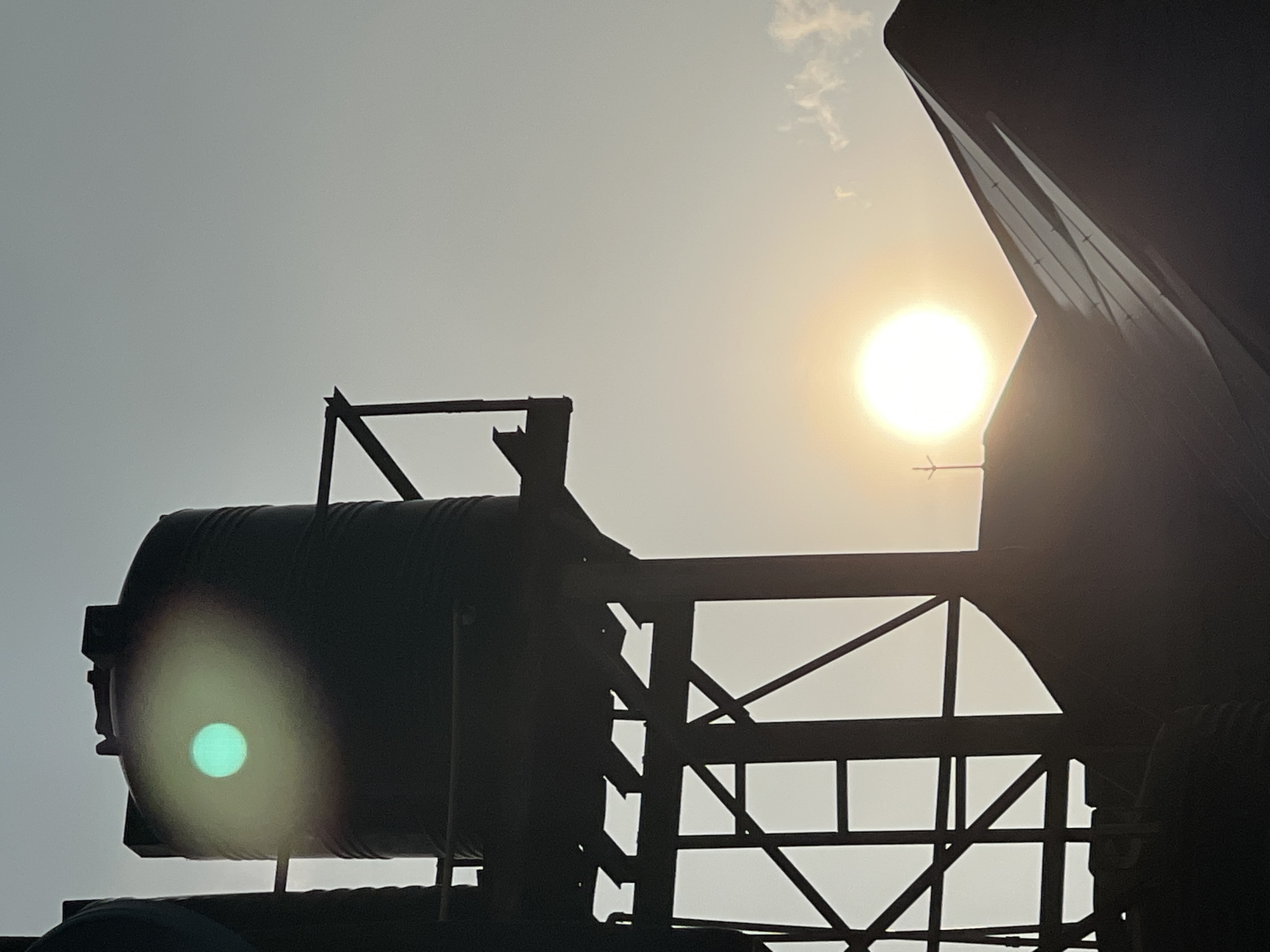 This is a vertical, medium shot photograph taken in Abuja, Nigeria, of a silhouetted water tank on a metal support structure against a hazy sky. The sun is visible as a bright, slightly diffused orb in the lower right quadrant of the image, casting a warm glow. A smaller, circular, greenish light artifact is present on the side of the water tank. A dark, angled roofline of a building is in the foreground, partially obscuring the base of the water tank structure. The overall impression is one of an urban setting at dusk or dawn.