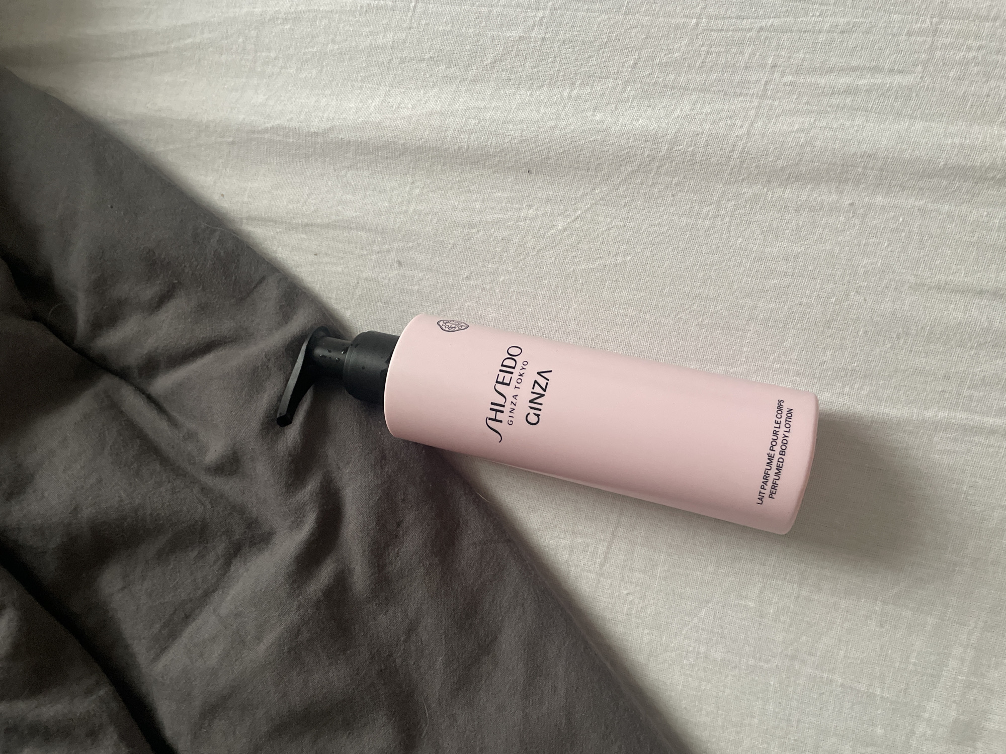 A pink Shiseido perfumed body lotion bottle with a black pump dispenser rests on a light-colored, textured surface, possibly linen. The bottle is positioned diagonally, with the top leaning towards the upper left. The label on the bottle reads "SHISEIDO GINZA TOKYO GINZA" and below that, in smaller text, "LAIT PARFUMÉ POUR LE CORPS PERFUMED BODY LOTION." To the left of the bottle, a dark grey, rumpled fabric is visible, creating a contrast in texture and color. The background is predominantly the light-colored surface, which appears to be a sheet or tablecloth, with visible wrinkles and texture. There are no people or animals present in the image. The scene is likely indoors, given the bedding-like textures. The location in Poroshkino, Russia is not discernible from the image content.