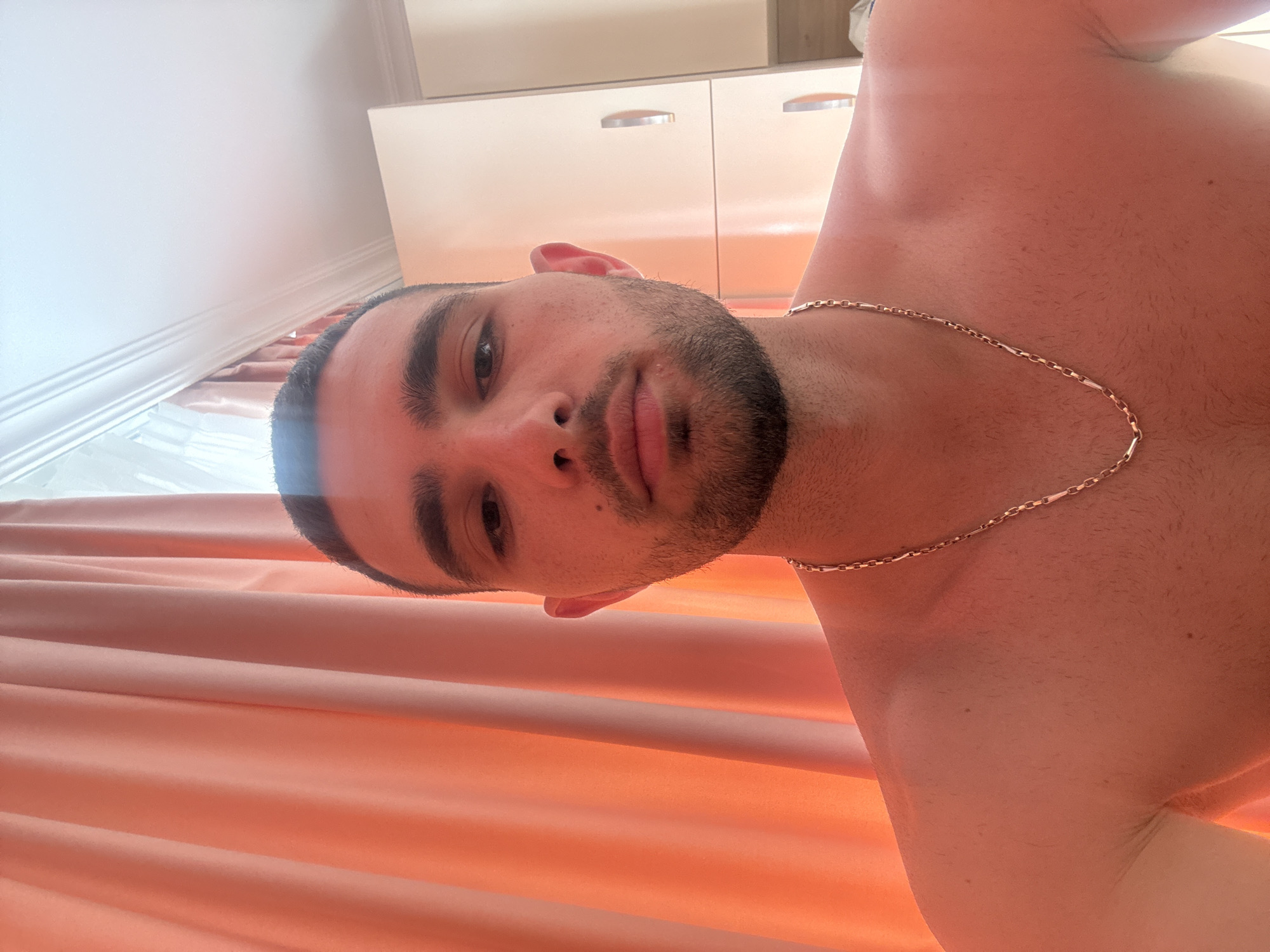 A close-up, chest-up selfie of a man is depicted. He is looking directly at the camera, with his head tilted slightly upwards. The man has dark hair, a beard, and is wearing a gold chain necklace. His skin is bare. In the background, there are peach-colored curtains on the left, and a white cabinet with two doors on the right. The ceiling, with decorative molding, is visible at the top. The lighting suggests an indoor setting, possibly a home. The overall impression is informal.