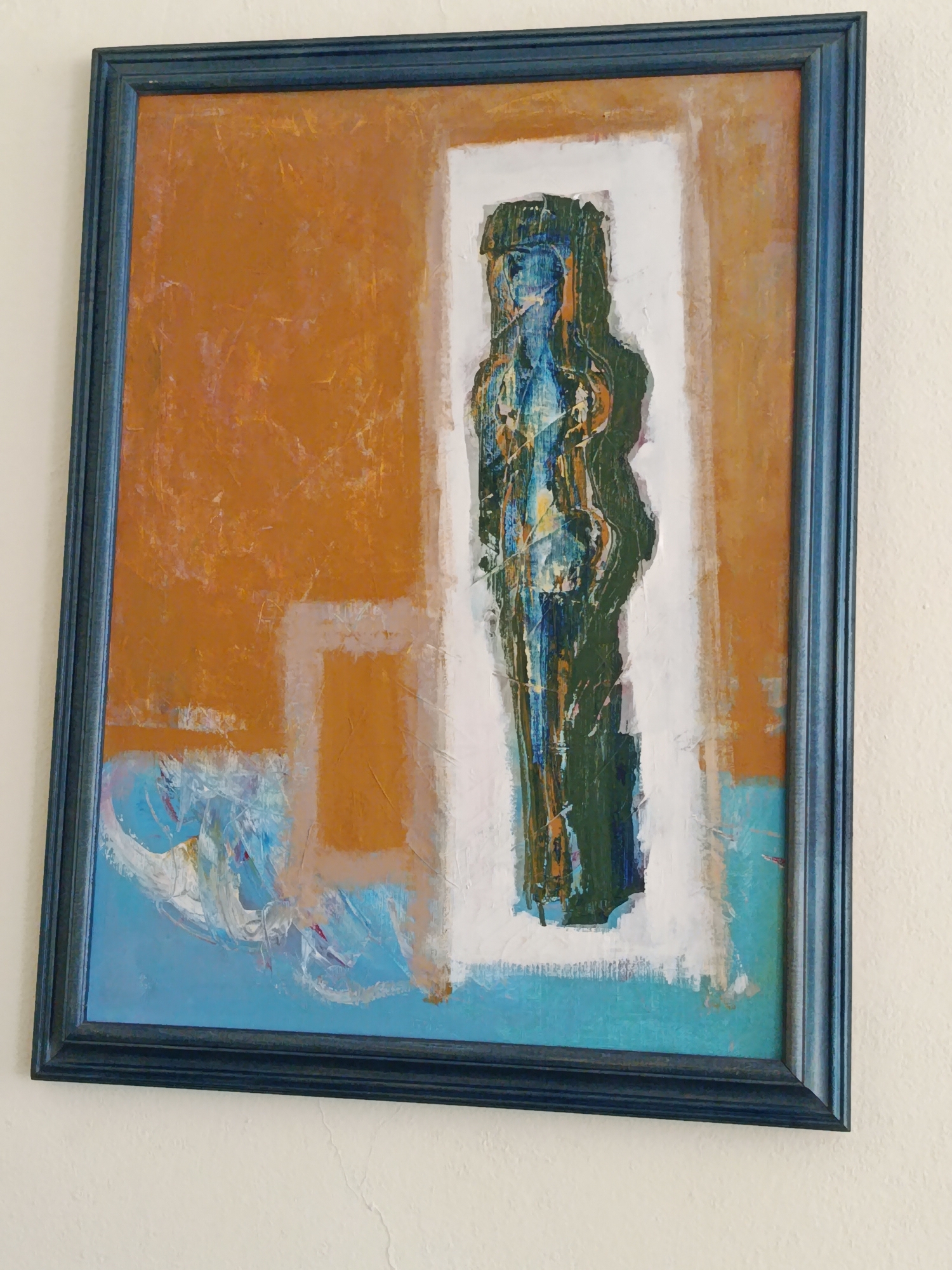 A framed abstract painting is hanging on a cream-colored wall. The painting is vertically oriented and features a dark blue, textured frame. The artwork itself is a mix of bold colors and textures. The background is divided into sections of warm orange and bright blue. A central white rectangular area contains a dark, vertical figure rendered in shades of green, blue, and yellow, with visible brushstrokes and textures suggesting a layered application of paint. The figure appears to be in silhouette, with some internal details showing through. The lower portion of the painting includes abstract swirls and strokes of blue and white paint, possibly representing water or movement. The overall impression is one of modern art, with an expressive and somewhat rough texture. The location is Varna, Bulgaria, with no specific identifiable landmarks visible in the image.