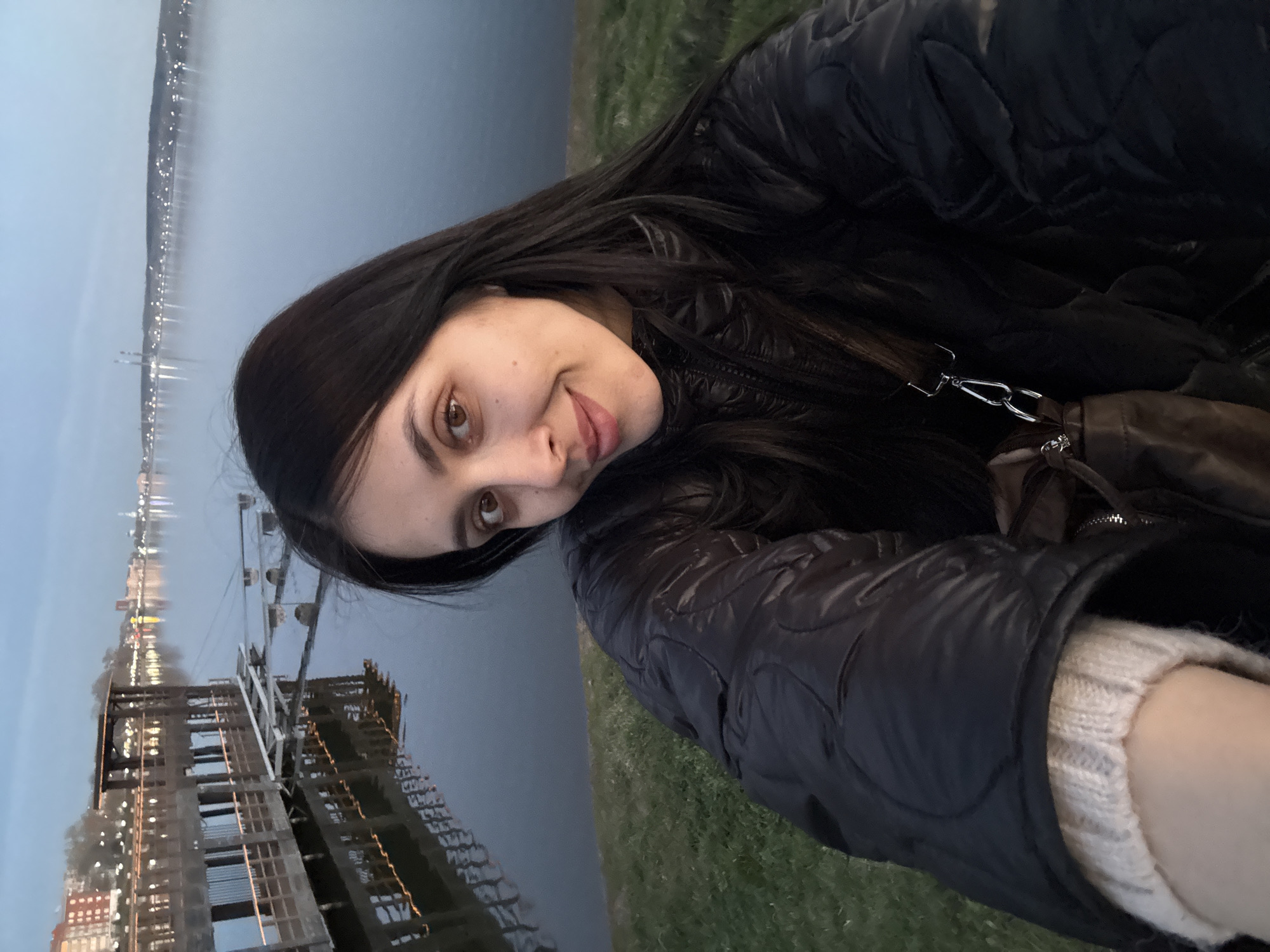 A selfie was taken of a woman with long black hair, wearing a black jacket and a cream-colored sweater. She is smirking slightly at the camera. A body of water is visible in the background, along with a building under construction. Distant city lights can be seen across the water. The grass is also visible in the foreground. Given the information, it is impossible to confirm this location is in Votkinsk, Russia.