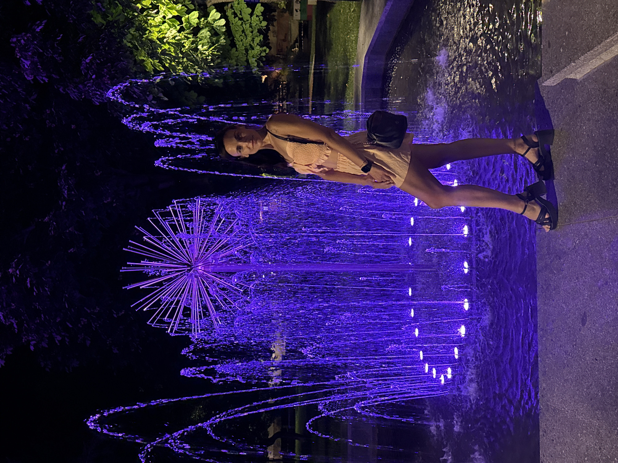 A woman stands in front of a fountain at night in Pleven, Bulgaria. The fountain has purple lighting, illuminating the water as it sprays upwards and outwards. The woman is wearing a light-colored, two-piece outfit and dark platform sandals. She has a dark crossbody bag slung over her shoulder. Behind the fountain, trees and foliage are visible, with some branches illuminated by the fountain's lights. The ground in front of the fountain is a paved walkway.