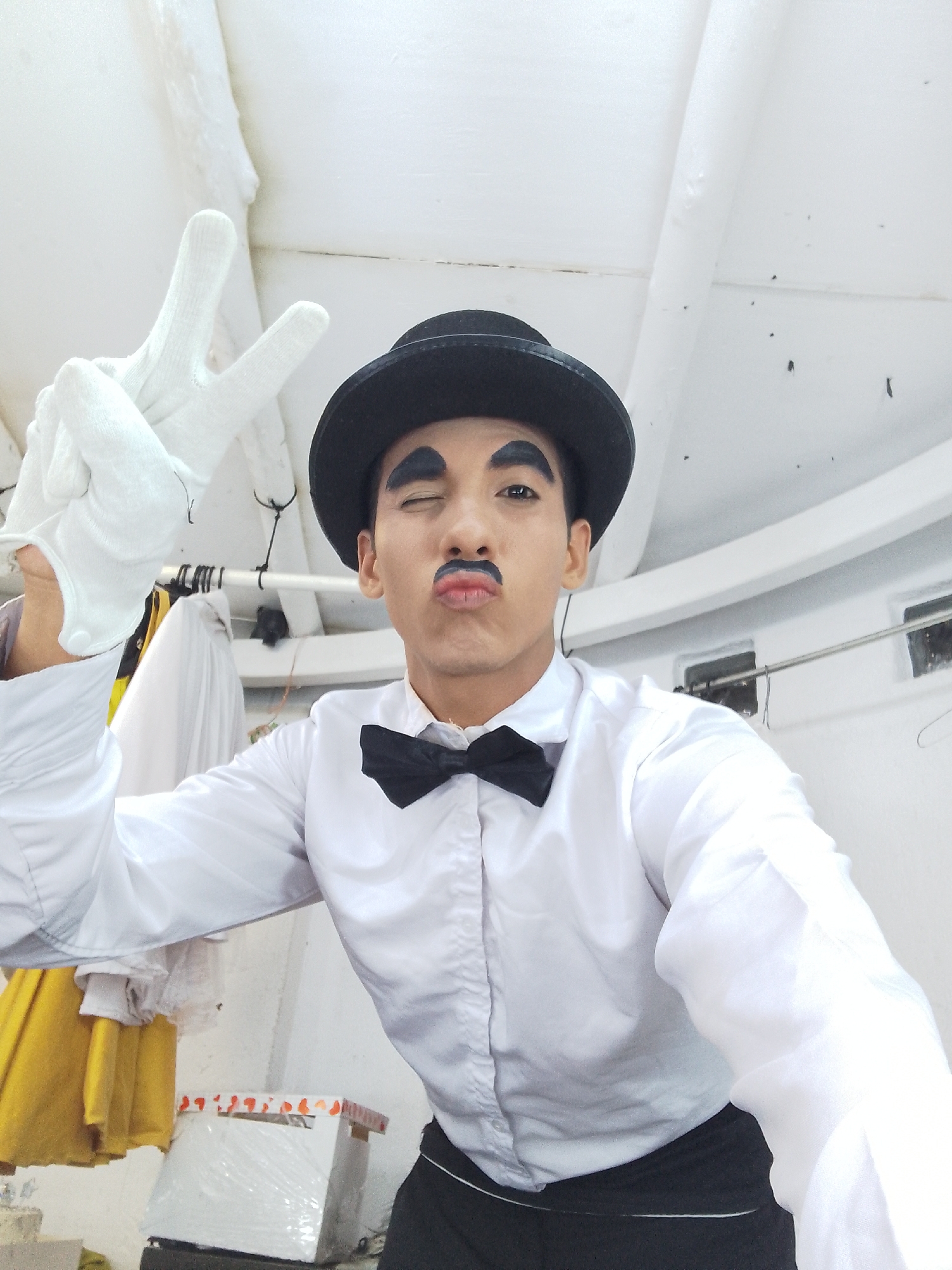 A person is dressed as Charlie Chaplin. They are wearing a black bowler hat, white gloves, a white dress shirt, a black bow tie, and a black skirt. They have drawn a black mustache on their face and are winking while making a peace sign. The setting appears to be indoors, with white walls and some articles of clothing hanging in the background. The location in the city of Unknown, Unknown is not known.