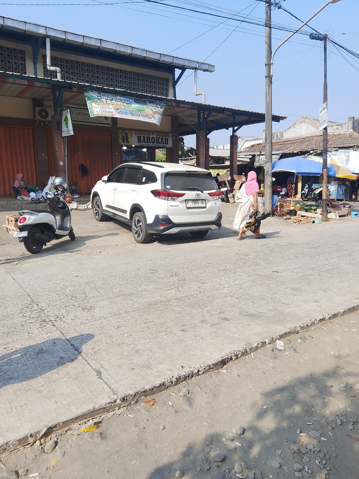 This is a medium shot, eye-level, outdoor photograph taken during the day in the city of Kalinyamatan, Indonesia. The image captures a street scene with a building, two vehicles, and two individuals. In the foreground, a light-colored concrete road surface dominates the lower half of the frame, with a dirt shoulder to the right. A shadow of a person is cast on the road.
To the left, a white scooter is parked facing right. Behind it, a white SUV is parked, facing left, angled slightly towards the viewer. The building in the background appears to be a market or shop with a corrugated metal roof supported by brown pillars. A banner advertising "ARTHA ABADI" and "SINERGI PASAR PLUS" is hung across the front of the building.
To the right of the SUV, a woman wearing a pink headscarf and a white dress is walking away from the viewer, carrying a dressed chicken in her right arm. She is wearing sandals. Behind her, under a blue umbrella, there appears to be a market stall with various items for sale, including what look like watermelons. The sky is clear and blue. The overall impression is of a busy marketplace on a sunny day.
