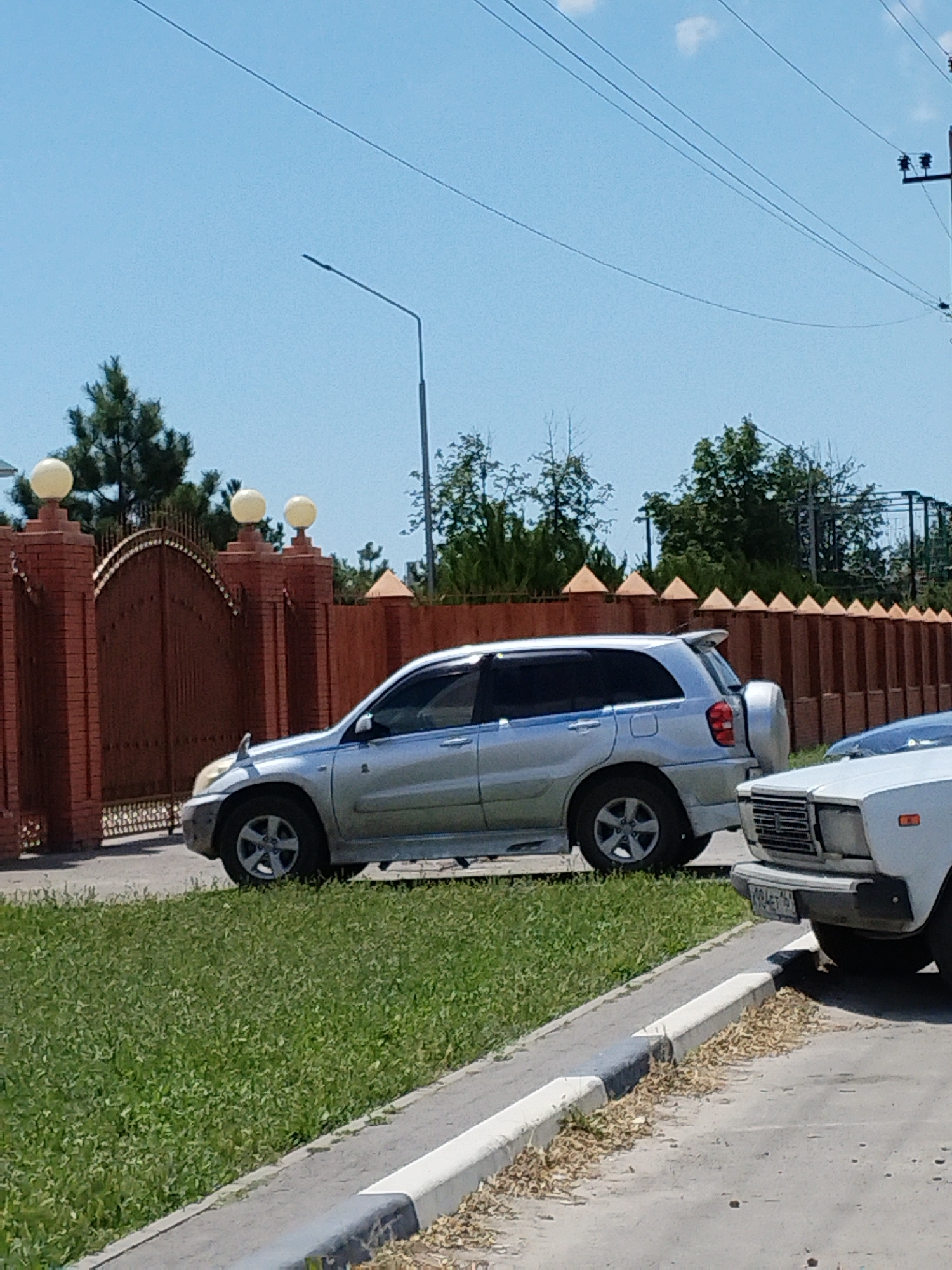 A silver Toyota RAV4 is parked on a grass verge next to a street in the city of Taganrog, Russia. The car is facing right. A white car is partially visible in the foreground, parked to the right of the RAV4. Behind the cars is a brick wall with a large arched gate. The gate is ornate and appears to be made of metal. The wall is topped with decorative elements and globe-shaped lights. In the background, there are trees and a blue sky with a few clouds. The scene appears to be a residential area.