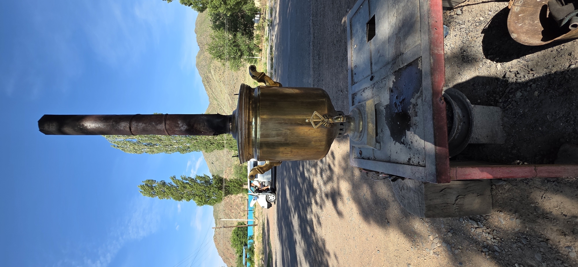 In Sasyk-Bulak, Kyrgyzstan, a brass samovar stands on a metal platform in the foreground. The samovar has two ornate handles and a spigot. A dark, cylindrical chimney extends upwards from the top of the samovar. The platform appears to be a makeshift structure with a red frame and a grey, perforated top. There are dark stains on the platform, suggesting recent use or liquid spillage.
Behind the samovar, a white van is parked on an asphalt road, with its hood open. A person is visible sitting in the driver's seat of the van. The ground beside the road is a mix of gravel and dirt. Tall, slender trees with green foliage stand in the middle ground. In the background, rolling hills with sparse vegetation and a clear blue sky with scattered wispy clouds complete the scene. The overall impression is an outdoor setting, possibly a roadside stop or a rural area.