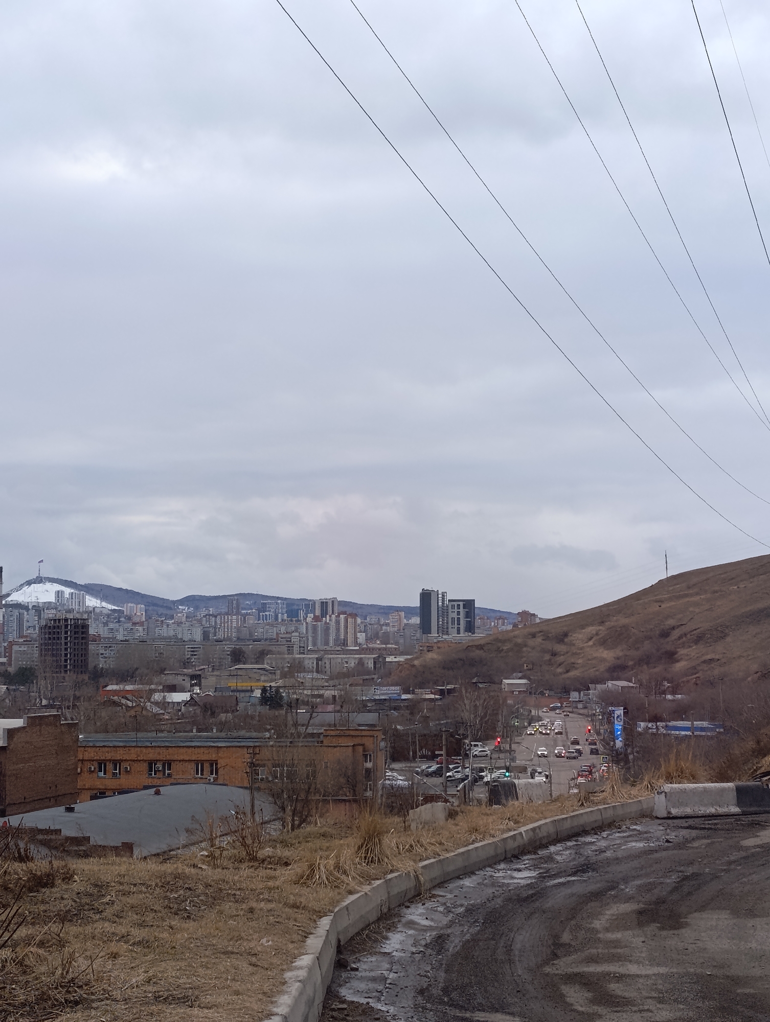 The image depicts an overcast day in Krasnoyarsk, Russia, with a view overlooking part of the city. In the background, buildings of varying heights make up the cityscape, including some modern high-rises and older, lower structures. A mountain partially covered in snow is visible in the distance with a structure on top. In the midground, a hillside slopes down to the right side of the frame. A road with cars and traffic lights passes along the base of the hill. In the foreground, there is a rough, dirty road and a low concrete barrier. The landscape is primarily brown and muted in color, with dried vegetation. Overhead, power lines traverse the sky.