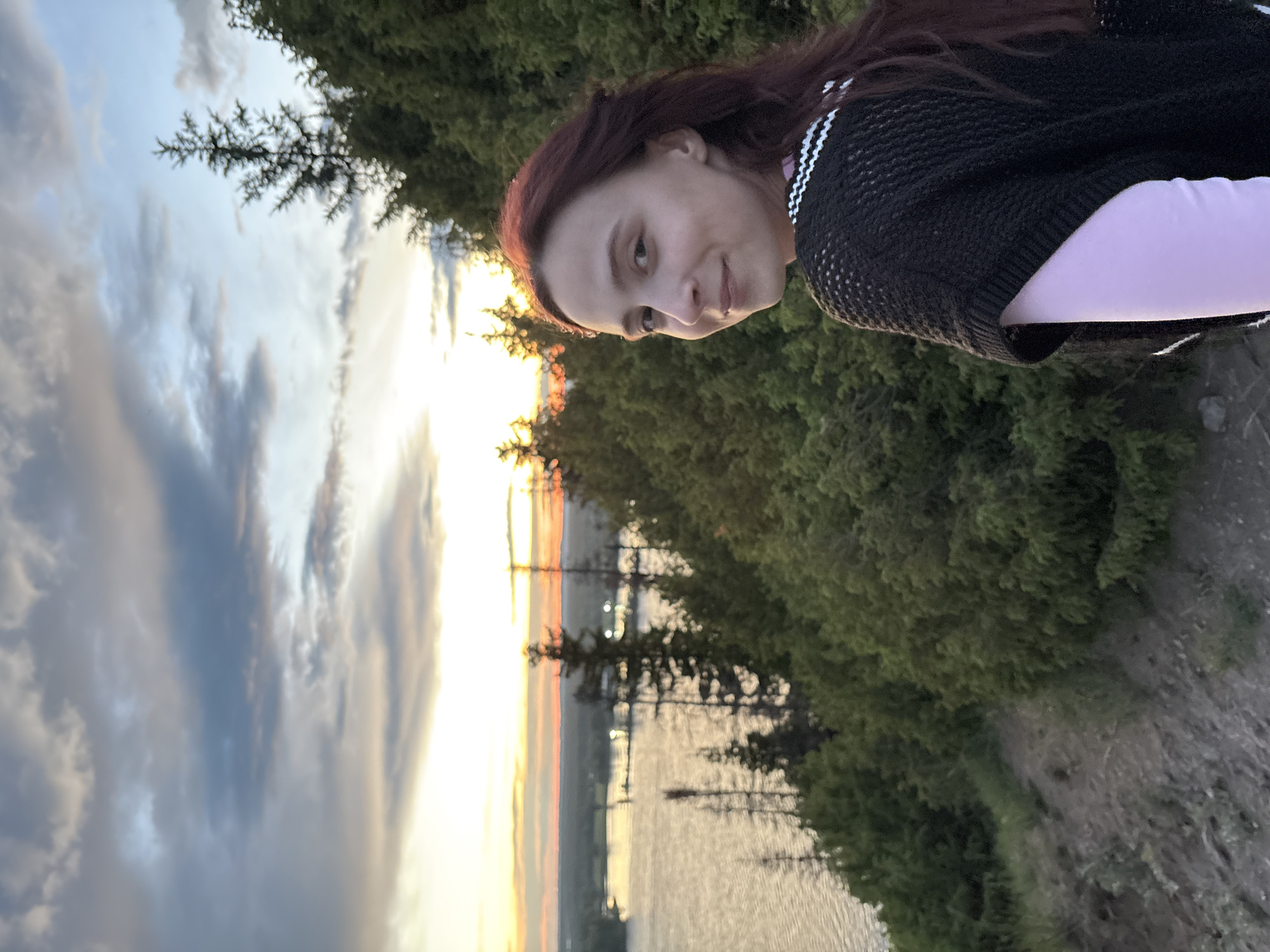 The image depicts a woman with auburn hair, wearing a black sweater over a pink shirt. She is looking at the camera with a slight smile. In the background, there is a view of a lake or river reflecting the colors of a sunset. The sky is partly cloudy, with hues of orange, yellow, and blue visible. Trees are visible on either side of the water. Based on the location provided (Ольховка, Russia), it is likely a natural setting near the water.