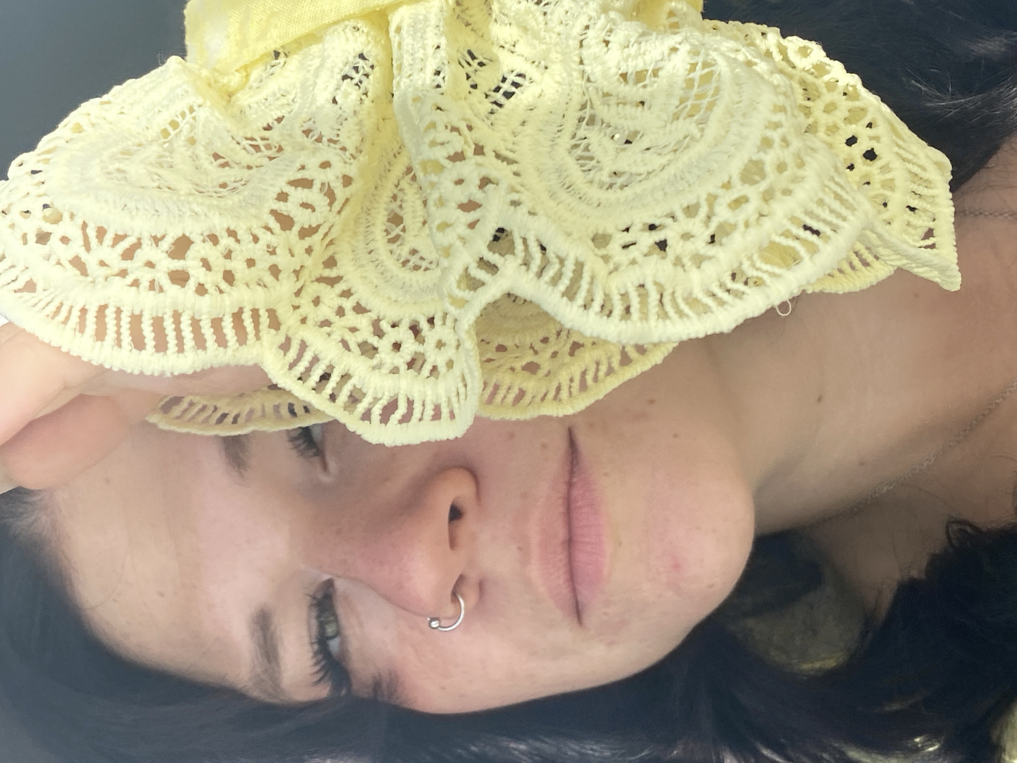 A close-up, slightly low-angle medium shot shows a young person with dark hair and light skin holding a yellow, lacy fabric to their face, obscuring the right side of their face. The person has a silver hoop piercing in their left nostril and appears to be looking towards the viewer with their left eye visible. The background is blurred and appears to be outdoors. There is no discernible indication of the city of Yanino-1, Russia in the image.