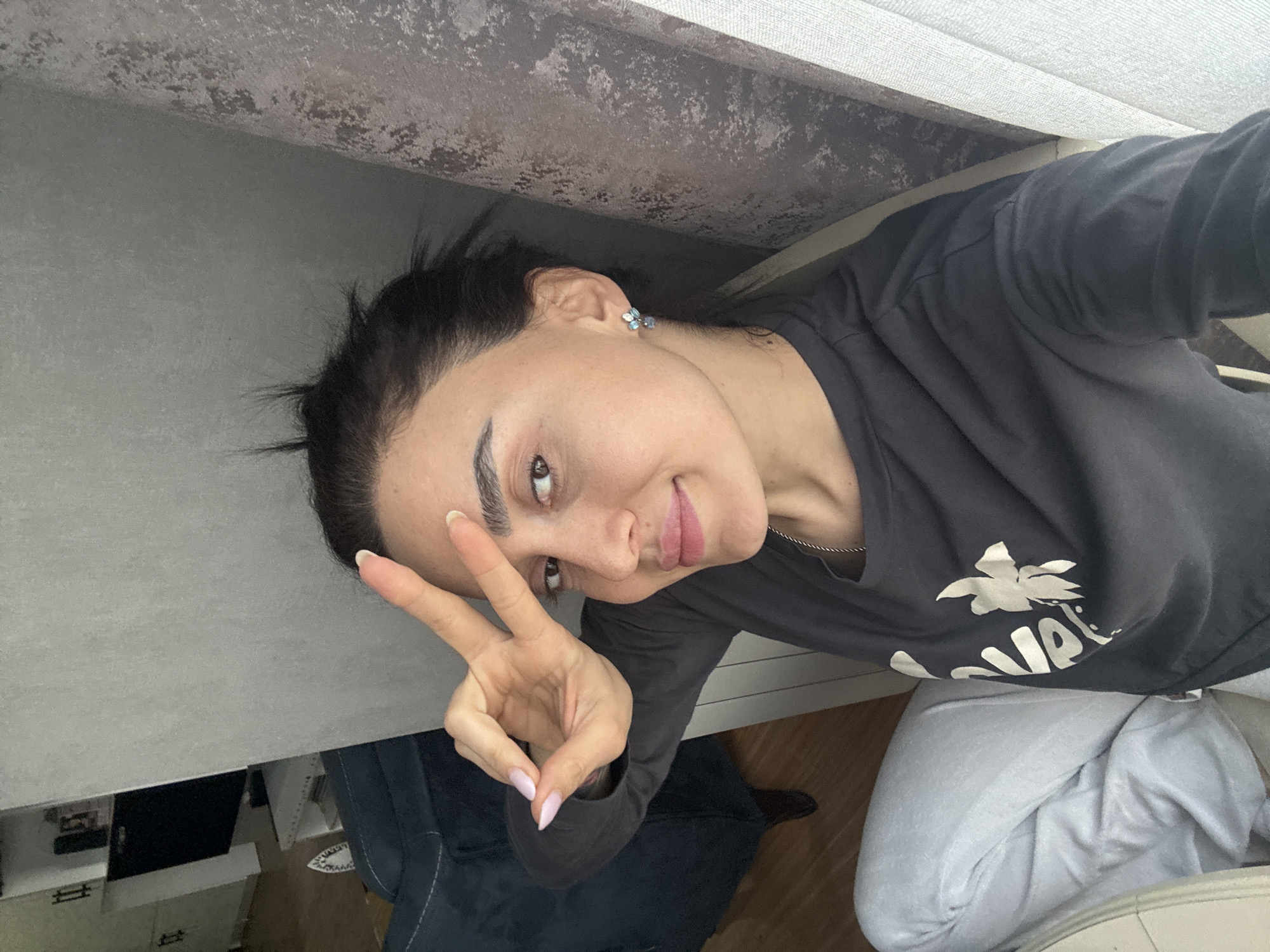 A woman is taking a selfie indoors. She is wearing a dark long-sleeved shirt with a white graphic on the front and light-colored pants. She is holding up her hand in a peace sign. Her hair is pulled back and she is wearing earrings. The background includes part of a wall, furniture, and flooring. It is not possible to determine the exact location in Tbilisi, Georgia from this image.