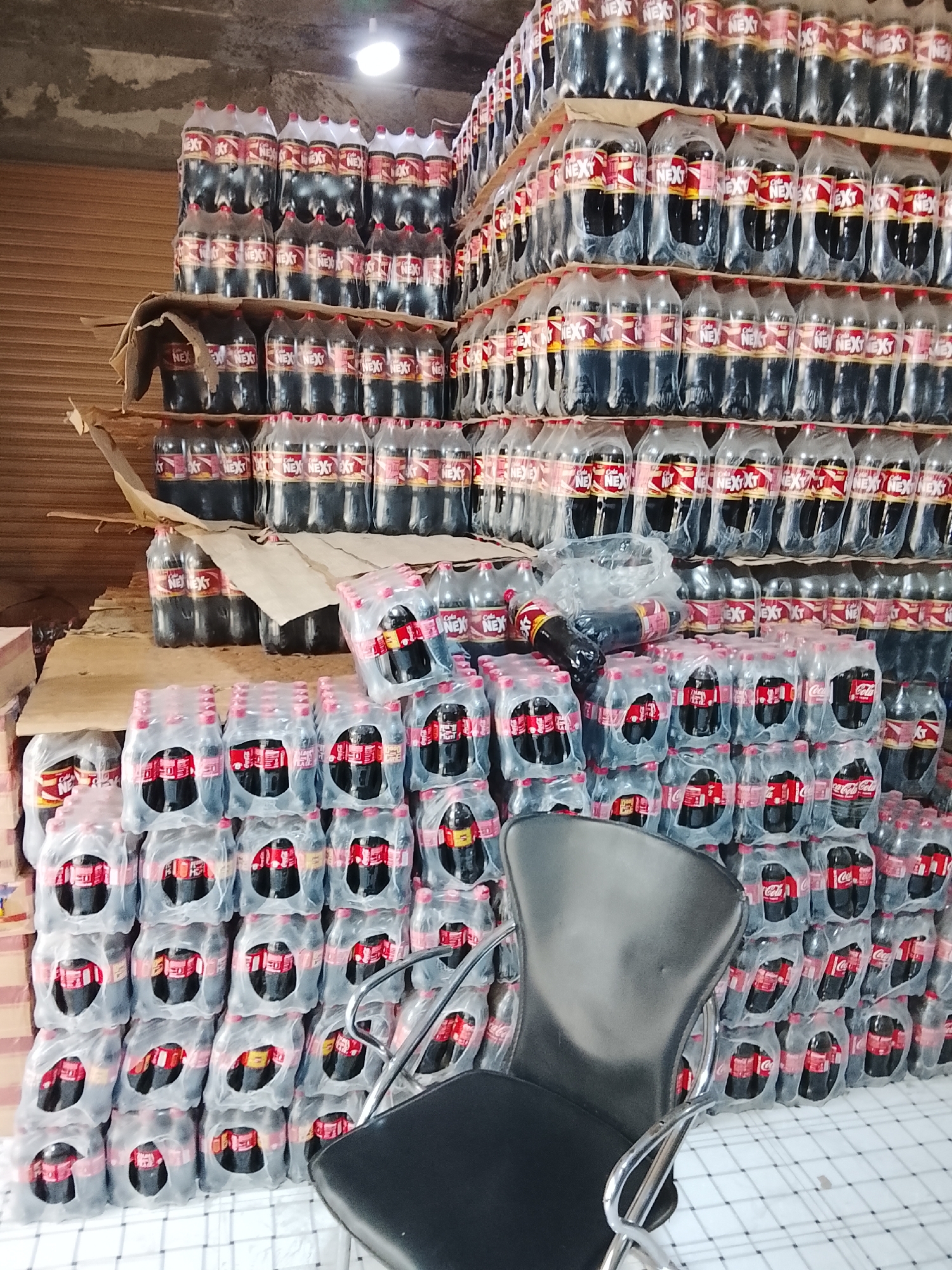 The image shows a large stock of "Next Cola" bottles, stacked high in plastic-wrapped packs. These are likely stored in a commercial space or warehouse. A black chair sits in front of the stacks. The location is suggested to be in Gulgasht Colony, Pakistan.