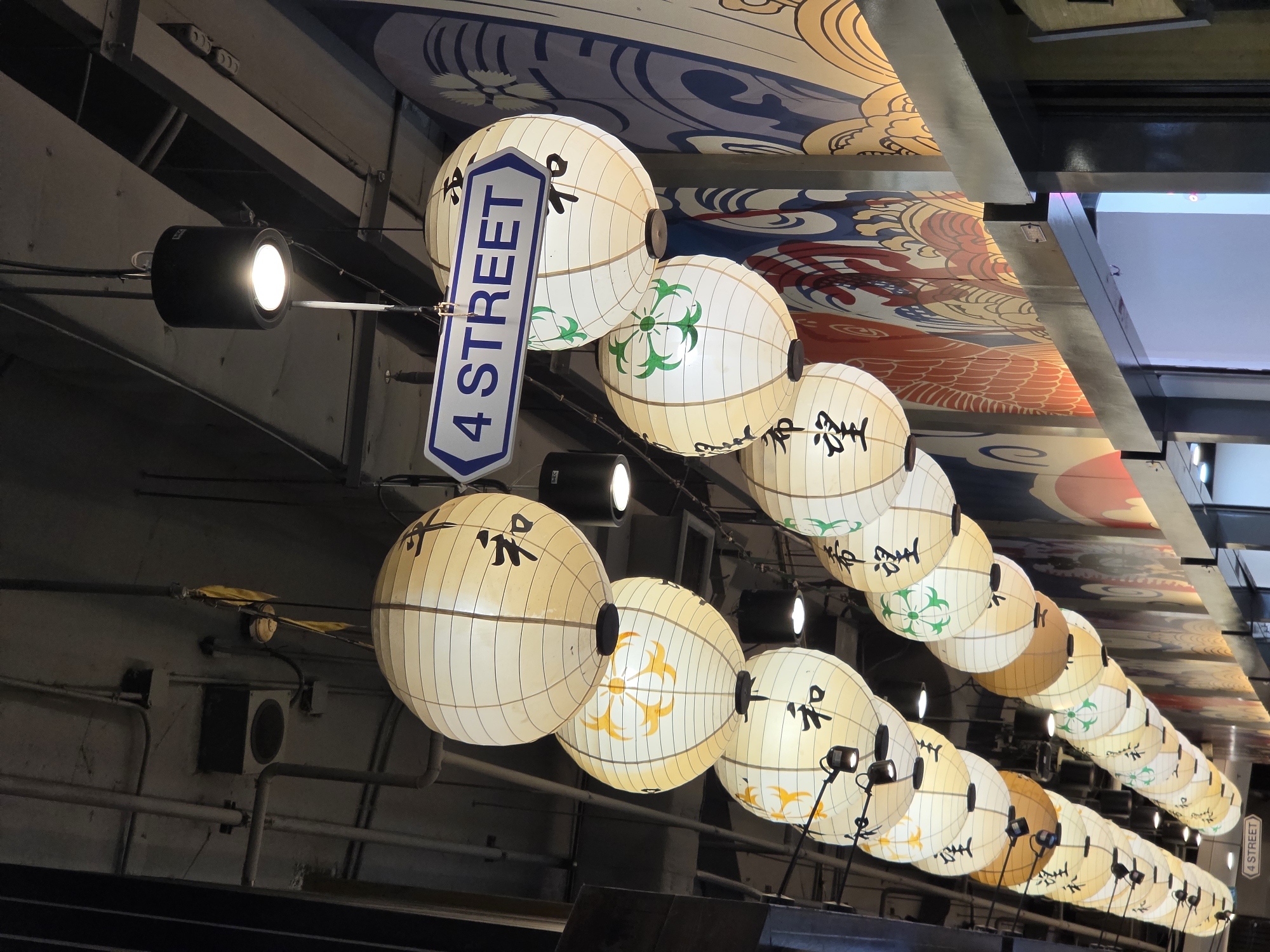 The image captures a vibrant indoor or covered walkway adorned with a long series of hanging spherical lanterns, hinting at an East Asian cultural theme, consistent with the Bangkok, Thailand context.
A prominent blue and white sign, shaped like an elongated hexagon, hangs near the foreground, clearly displaying "4 STREET" in capital letters. The numerous lanterns, primarily off-white or cream-colored, feature various designs: many are decorated with bold black Japanese or Chinese characters (likely auspicious symbols), while others display green floral patterns or yellow-orange star-like motifs. These lanterns are illuminated by multiple black spotlights mounted on overhead beams, some of which are brightly lit, casting a warm glow.
Above the lanterns, a richly decorated ceiling or canopy is visible, painted with traditional East Asian-inspired artwork. This artwork includes swirling blue waves, sections resembling red or orange dragon scales, and ornate yellow or gold cloud and floral patterns. The infrastructure of the ceiling, including dark metal beams, conduits, and wires, is also visible. On the upper left, part of a wall shows a mounted speaker or vent.
The scene suggests an evening or indoor setting where artificial lighting creates a festive and decorative atmosphere. No people or specific activities are depicted, focusing instead on the elaborate cultural decor.