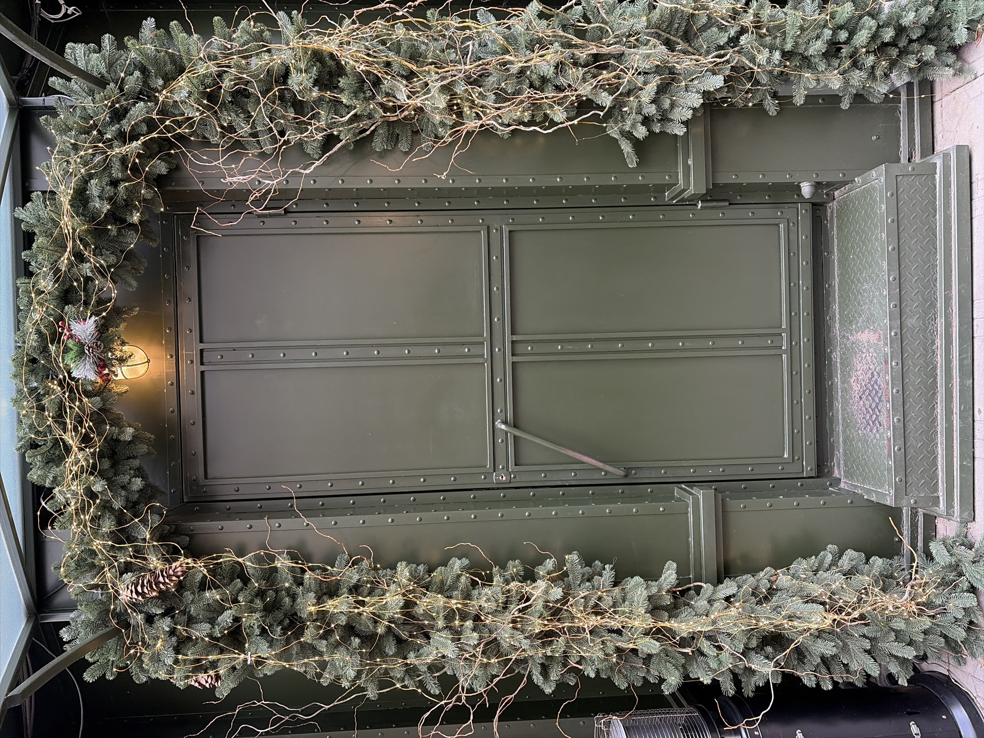 A low-angle shot depicts a green, riveted, rectangular structure adorned with a garland of evergreen branches, bare twigs, and small, warm-toned lights. The garland frames the structure on three sides. On the left side of the frame, a decorative element including pine cones and red berries is visible. To the right of the rectangular structure, a set of diamond-patterned metal steps is seen. The style and composition of the image suggest a festive or decorative setting. The exact location in Sofia, Bulgaria, cannot be determined from the given image.