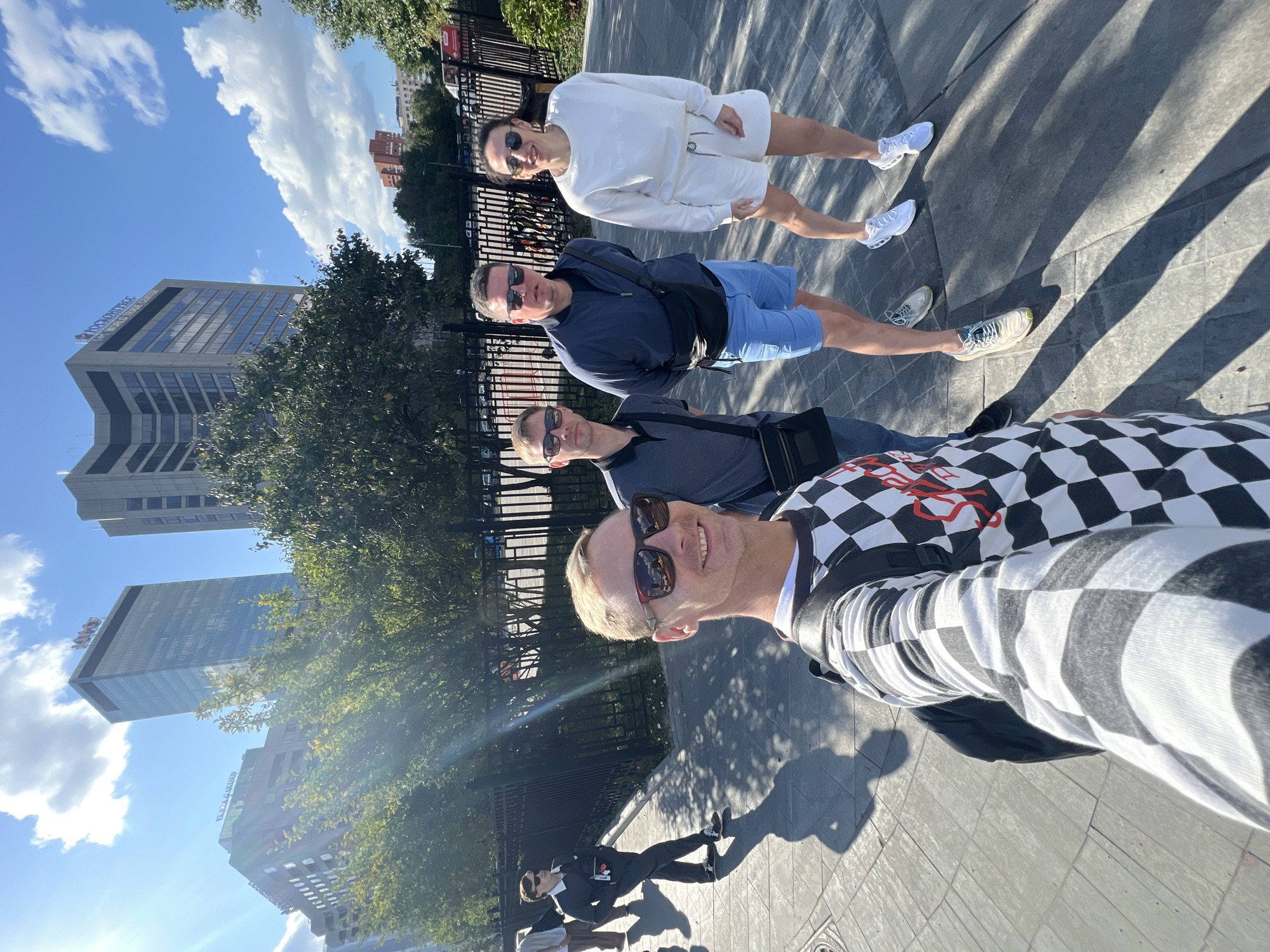 The image shows four individuals standing on a sidewalk in Moscow, Russia. The sun is shining brightly, casting long shadows on the ground. In the background, there are several tall buildings, including the World Trade Center Moscow. The individuals appear to be friends, and they are all smiling and looking at the camera. The person in the foreground is taking a selfie. The other three people are standing behind him, also posing for the photo. The scene suggests a casual outing on a sunny day in the city.