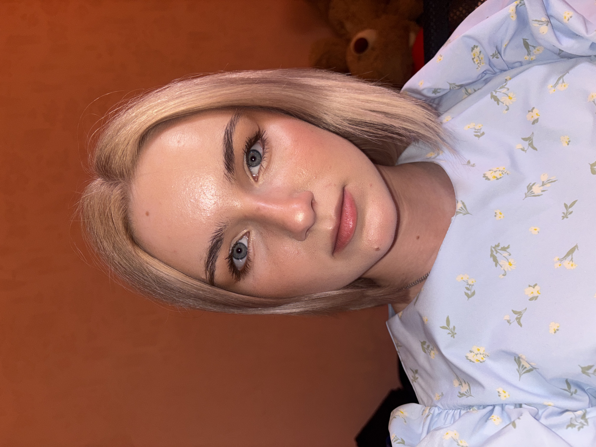 A close-up portrait, taken indoors in what is likely Saint Petersburg, Russia, features a young woman with fair skin, short blonde hair, and prominent blue eyes. She looks directly at the camera with a neutral, calm expression. Her eyebrows are dark and well-defined, and her lips are full and light pink. A small mole is visible on her right cheek.
She wears a light blue top or dress featuring a delicate pattern of small white and yellow flowers with green leaves. The garment includes ruffled details around the neckline or sleeves. A thin, silver-colored chain necklace is visible around her neck.
The background is a solid, warm reddish-brown, possibly a wall or curtain, indicating an indoor setting. A blurred, dark brown teddy bear is partially visible in the upper right, contributing to a personal and cozy atmosphere, perhaps a bedroom. The lighting is warm and artificial, highlighting her features. There is no visible text in the image.